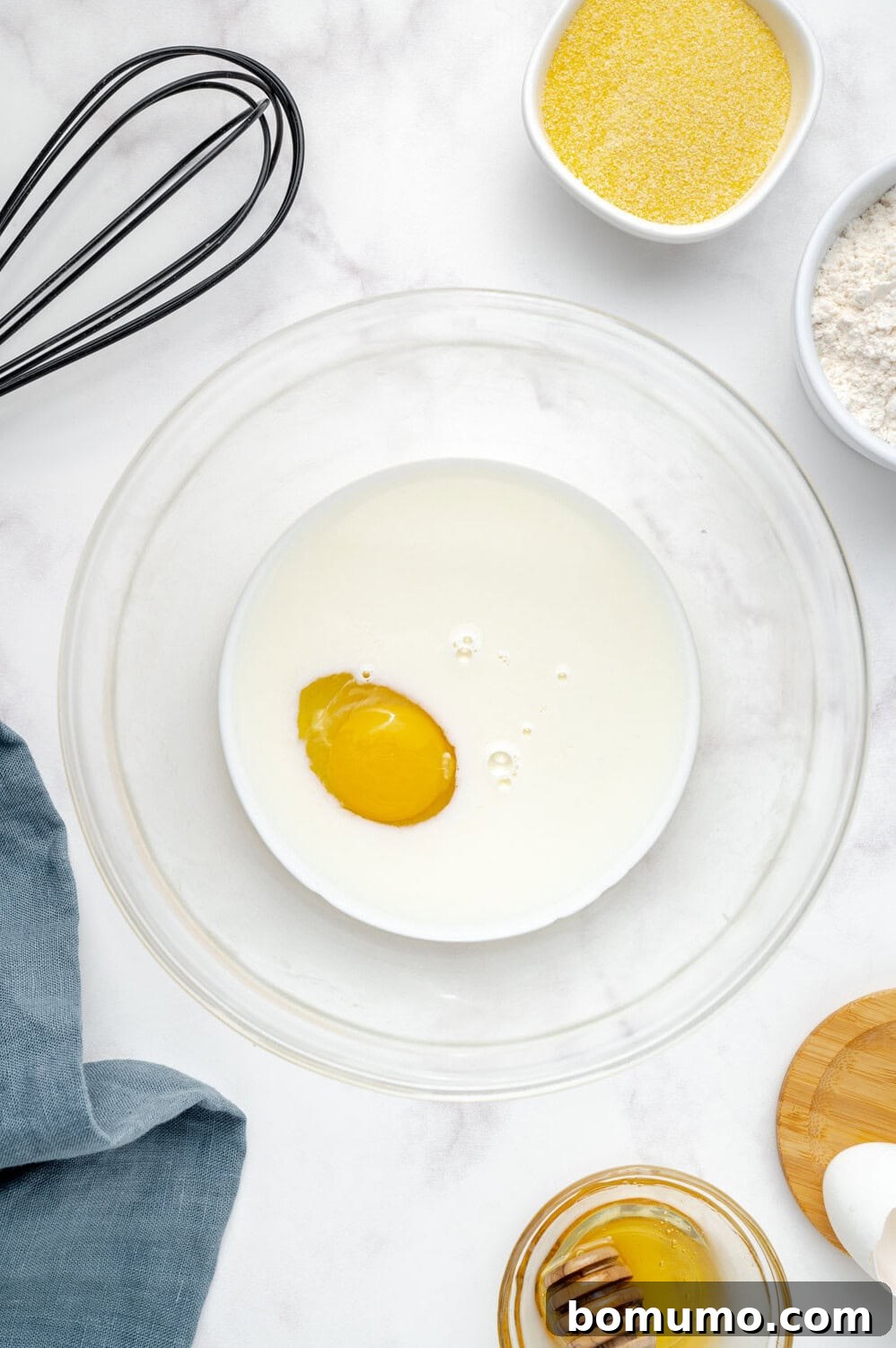 A clear mixing bowl contains milk and a vibrant egg yolk, ready to be combined. Nearby, a whisk awaits, alongside separate bowls of all-purpose flour and golden cornmeal, a delicate dish of honey, and a soft blue cloth, setting the scene for baking.