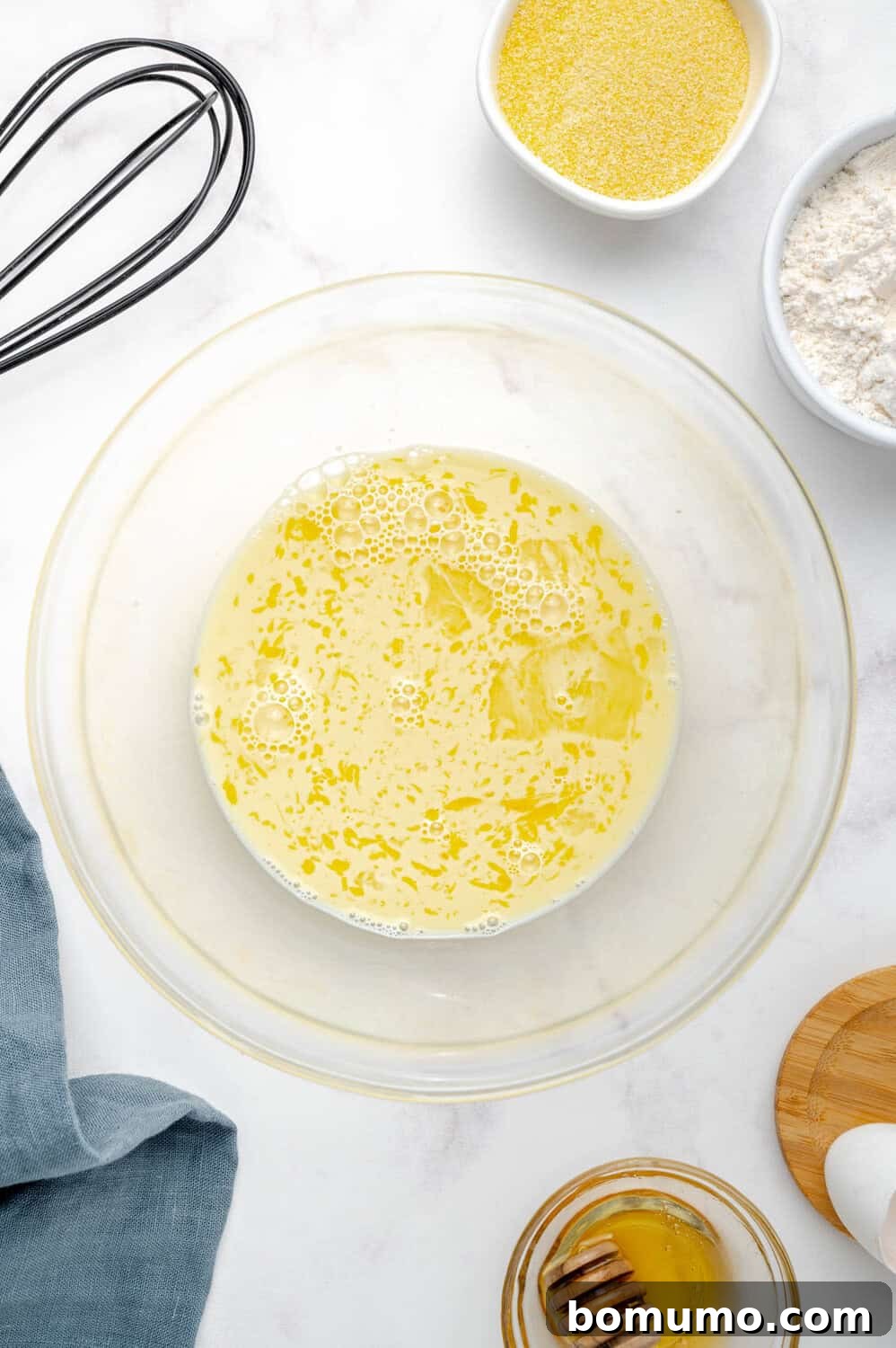 A mixing bowl filled with perfectly combined wet ingredients, including a glistening layer of oil, rests on a pristine white countertop. Surrounding it are a whisk, separate bowls of flour and cornmeal, a soft cloth, a jar of honey, and a wooden board, all prepared for the next step in the recipe.