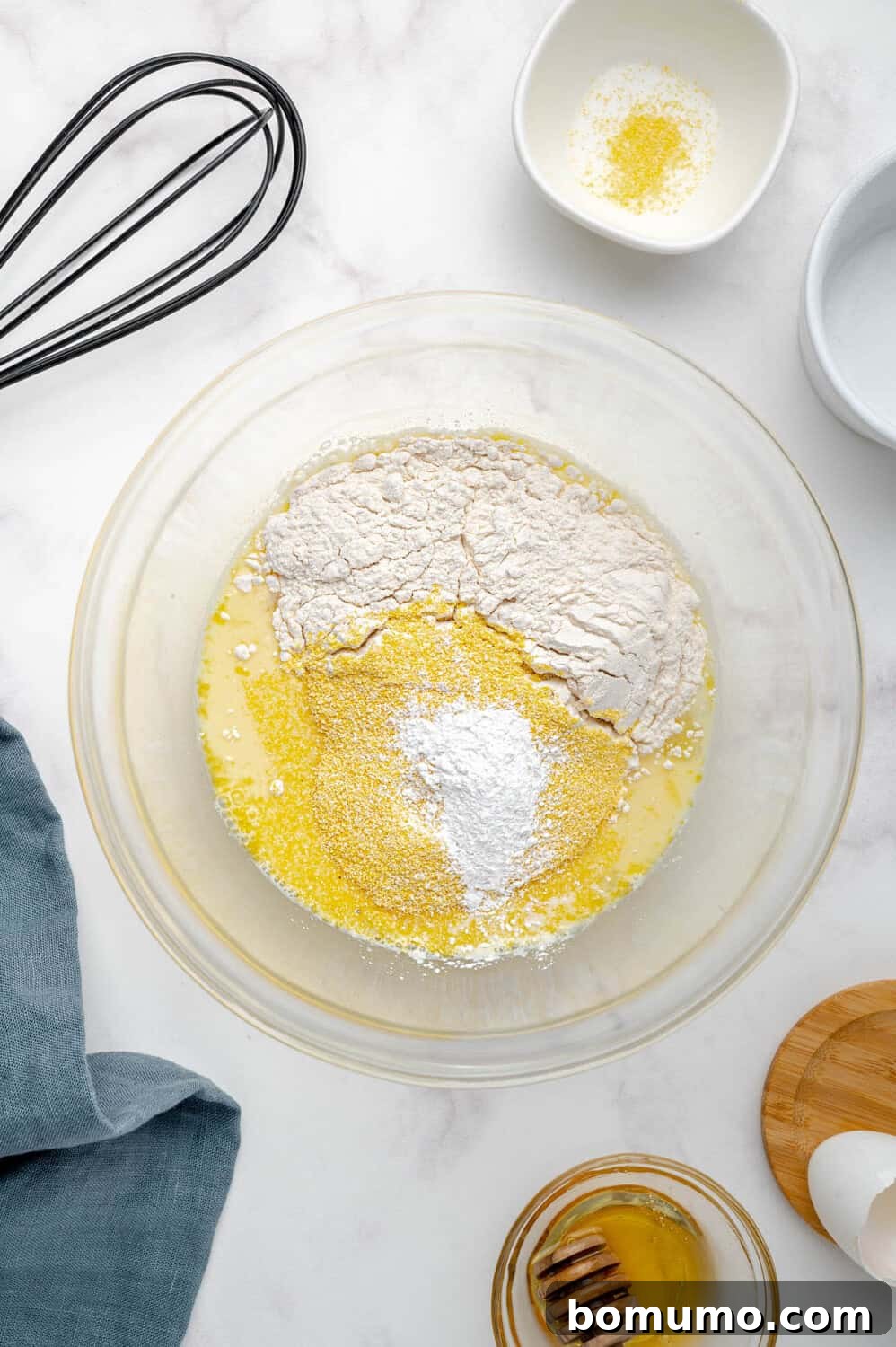 A mixing bowl filled with the dry ingredients for cornbread pancakes, including flour, cornmeal, and baking powder, expertly layered over the wet mixture. A whisk, a soft cloth, a jar of honey, and small prep dishes are arranged neatly on a marble countertop, ready for the final stir.