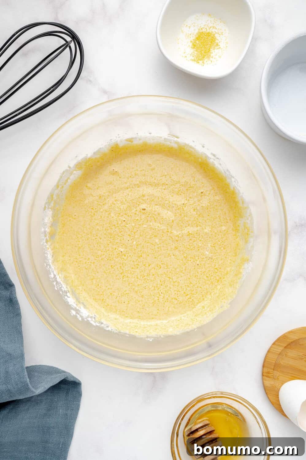 A mixing bowl contains a vibrant yellow batter, perfectly combined for cornbread pancakes. Alongside, a whisk, a jar of honey, and small bowls of salt and flour rest on a sleek marble countertop, showcasing the harmony of preparation.