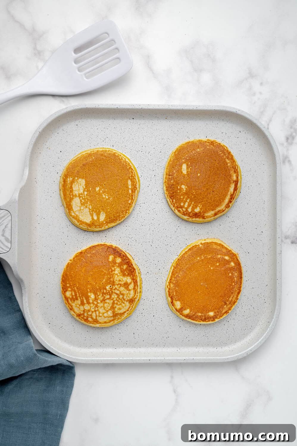 Four perfectly golden-brown cornbread pancakes sizzle on a pristine white griddle, radiating warmth. A reliable gray spatula and a neatly folded blue napkin rest nearby on a cool marble surface, ready for service.