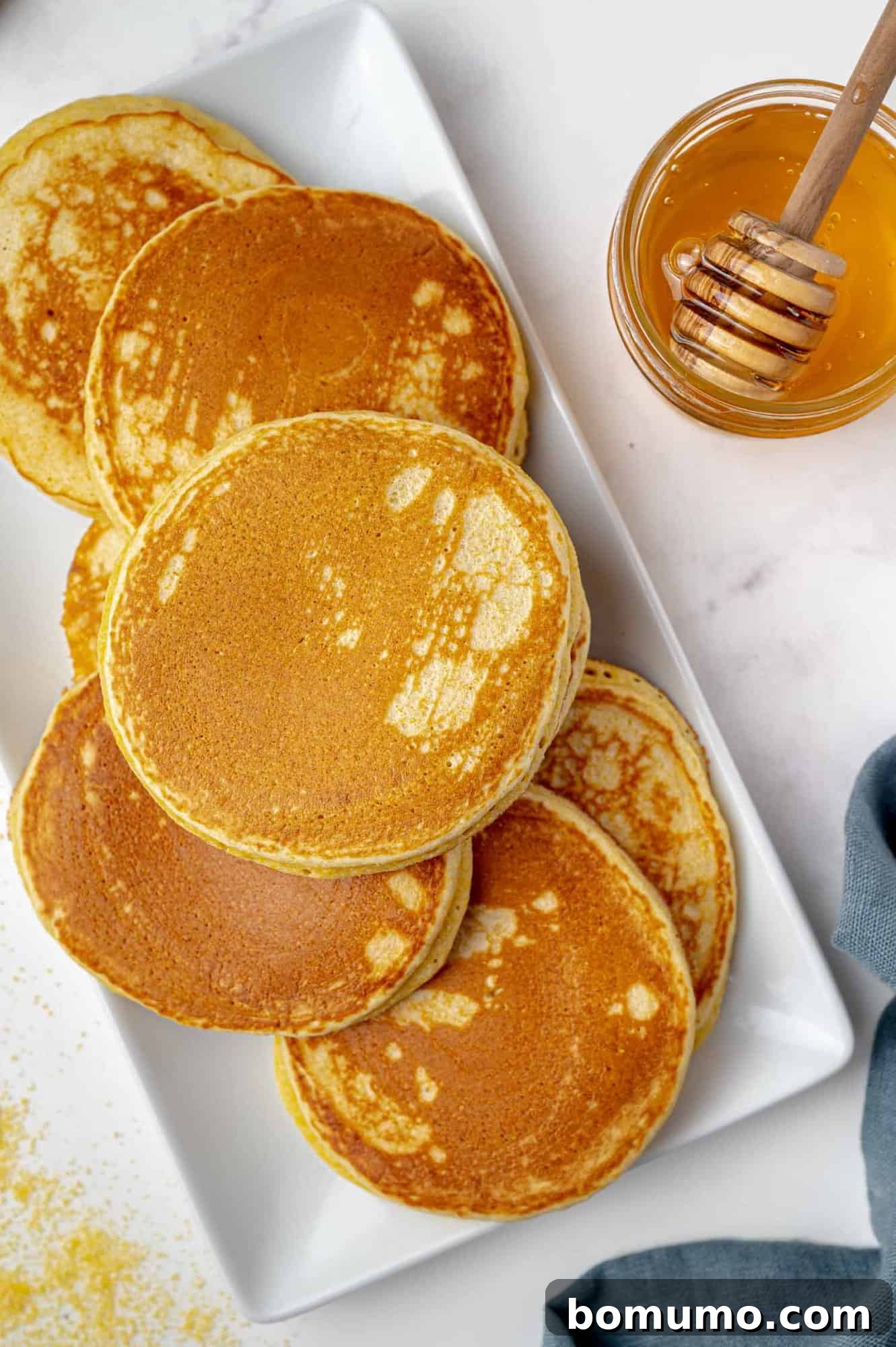 A neatly stacked tower of golden cornbread pancakes rests on a crisp white rectangular plate, exuding a comforting aroma. Beside it, a rustic jar of honey awaits, complete with a wooden honey dipper, promising a touch of natural sweetness.