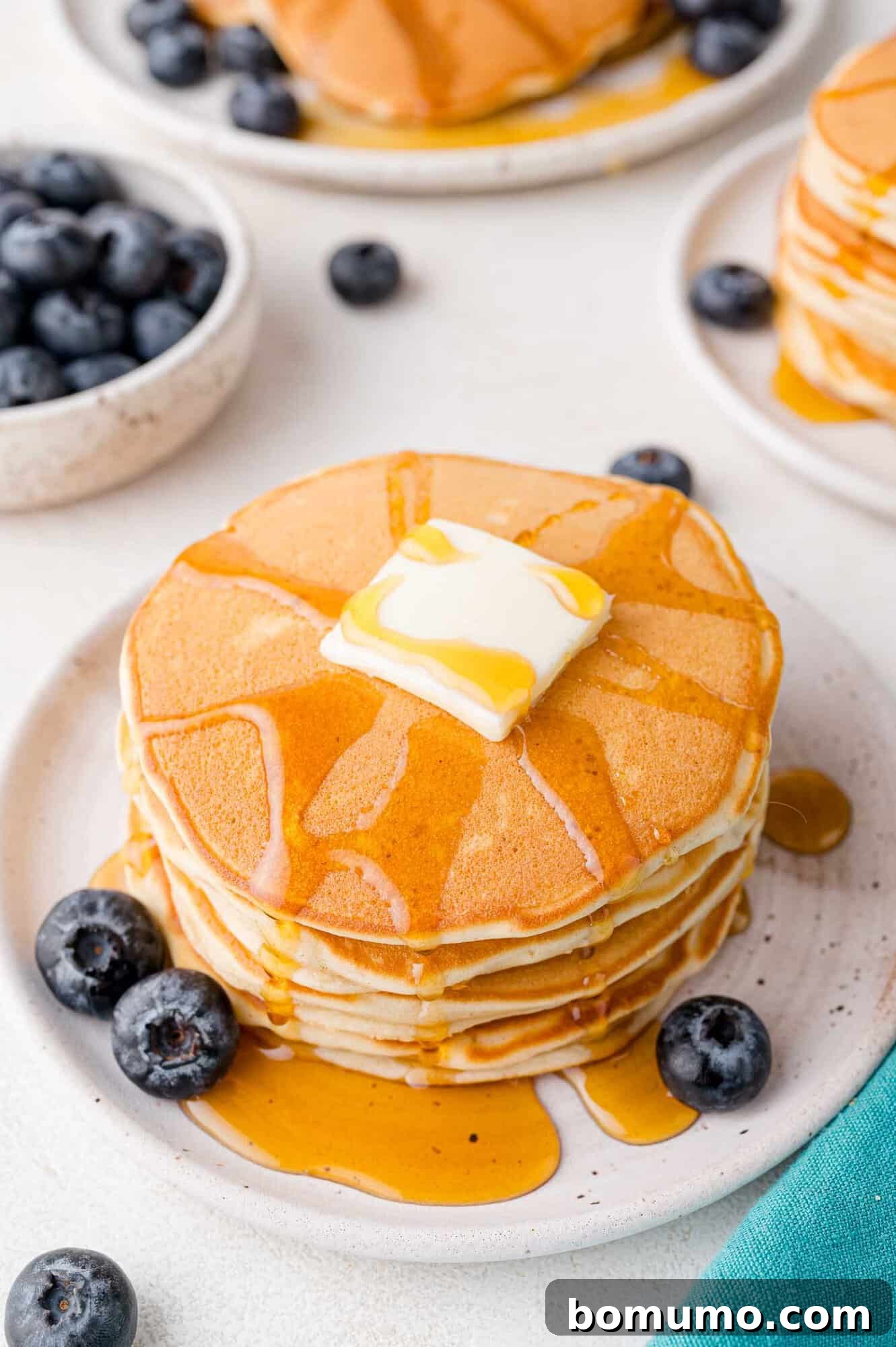 A stack of golden-brown sweet cream pancakes, generously topped with a pat of melting butter and glistening syrup, surrounded by fresh blueberries on a pristine white plate.