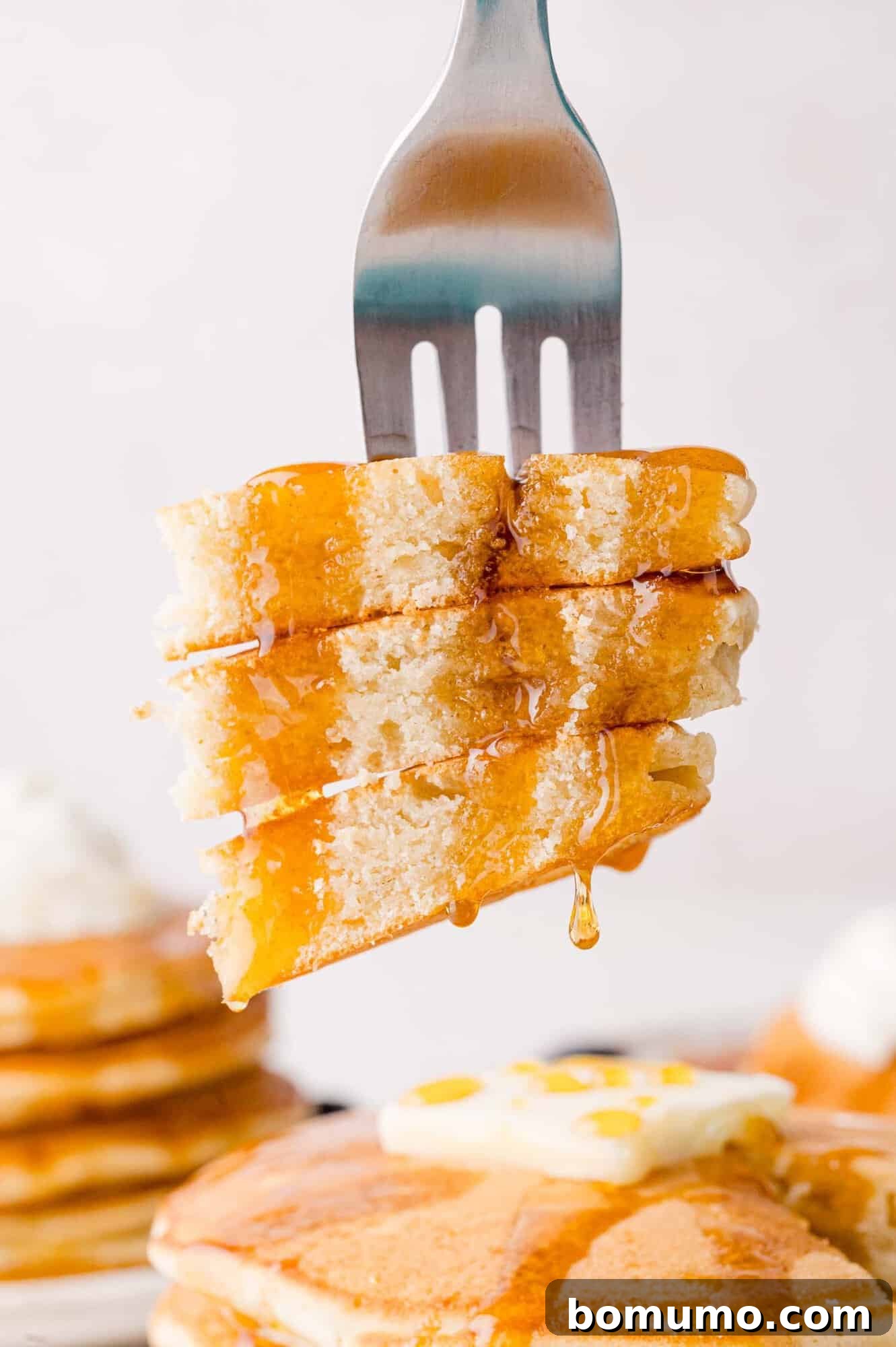 A fork piercing a stack of syrup-drizzled sweet cream pancakes, with a blurred background showing more pancakes topped with butter.