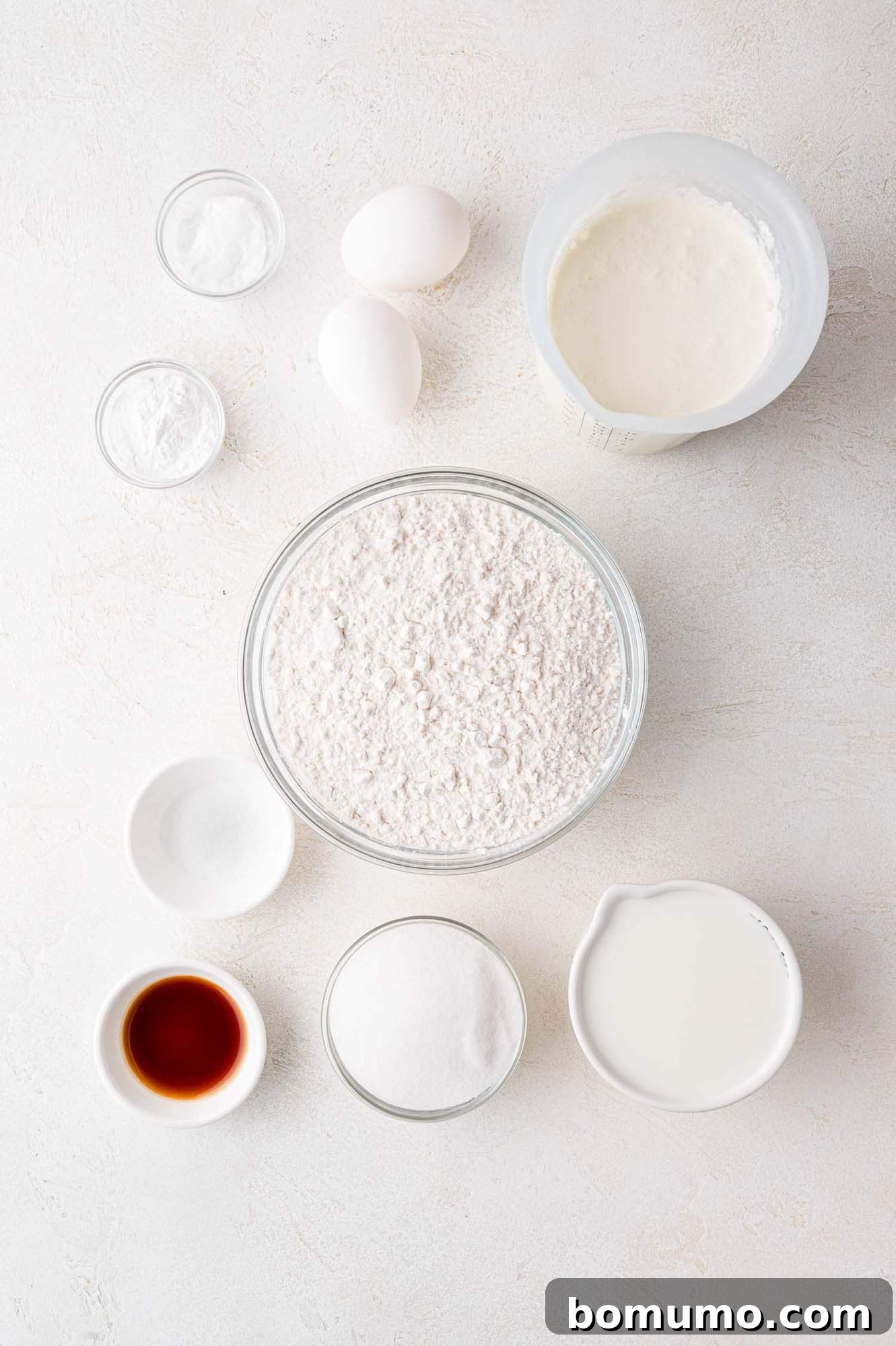 Various baking ingredients artfully arranged on a rustic wooden table, including bowls of flour, sugar, baking soda, and baking powder, alongside a jug of milk, vanilla extract, and fresh eggs.