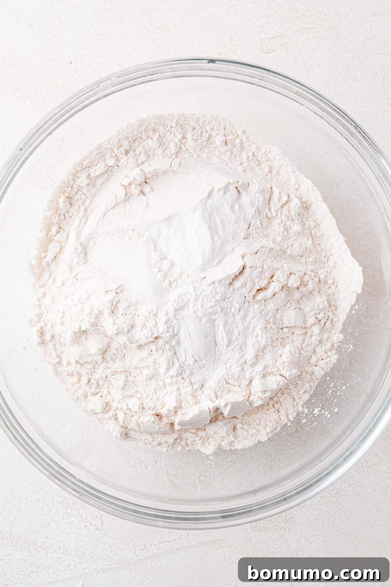 A clear glass bowl filled with finely sifted all-purpose flour sits prominently on a clean, light-colored kitchen surface, ready for the next step in the baking process.