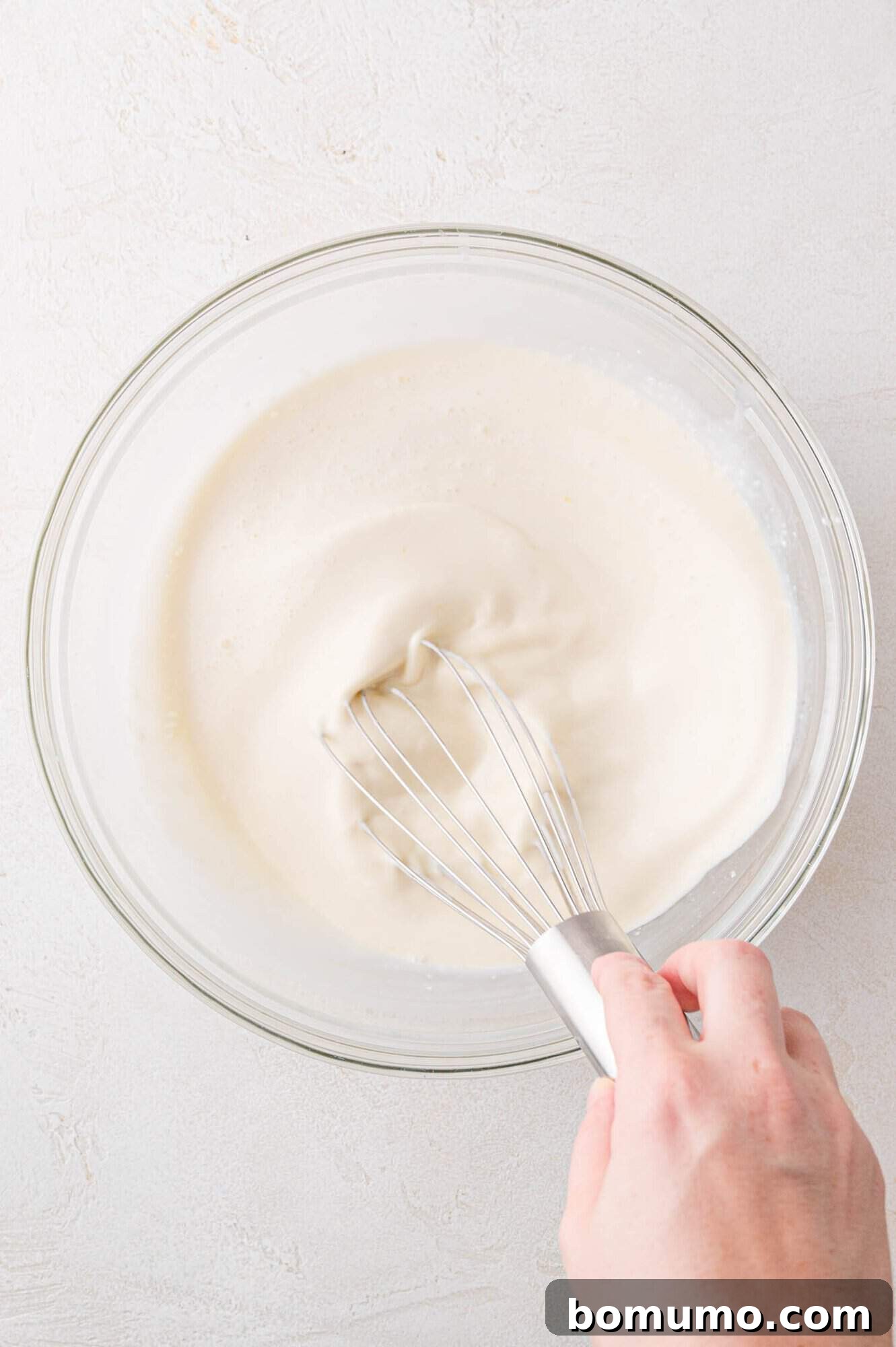 A close-up shot of a hand expertly whisking a smooth, creamy white batter in a transparent glass bowl, set against a pristine light surface, highlighting the fluidity of the mixture.