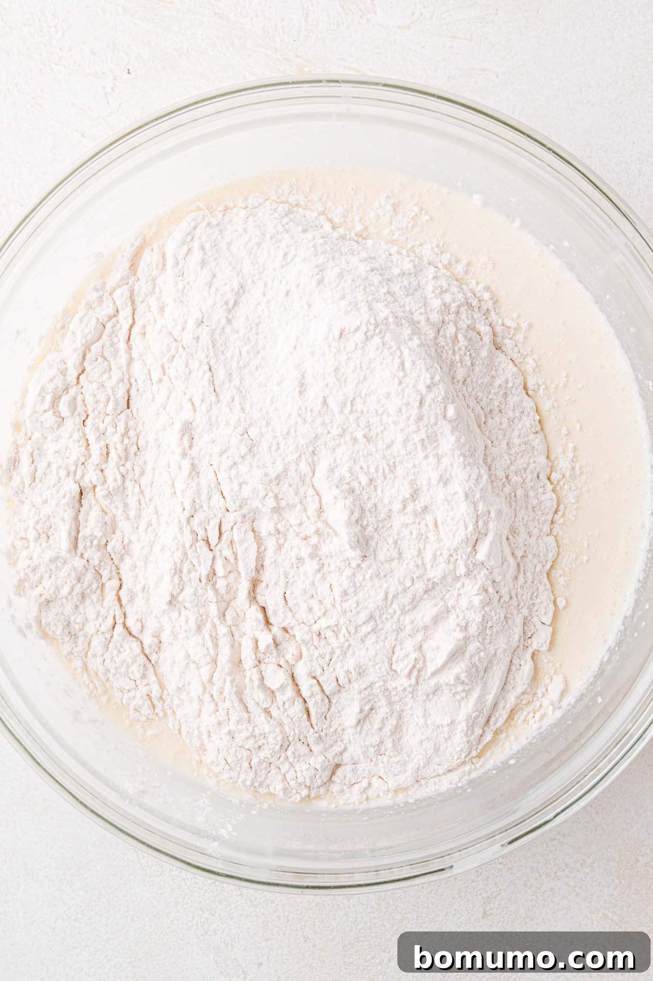 A transparent glass bowl with a mound of dry flour mixture carefully heaped onto a creamy liquid base, illustrating the moment before combining the ingredients for pancake batter.
