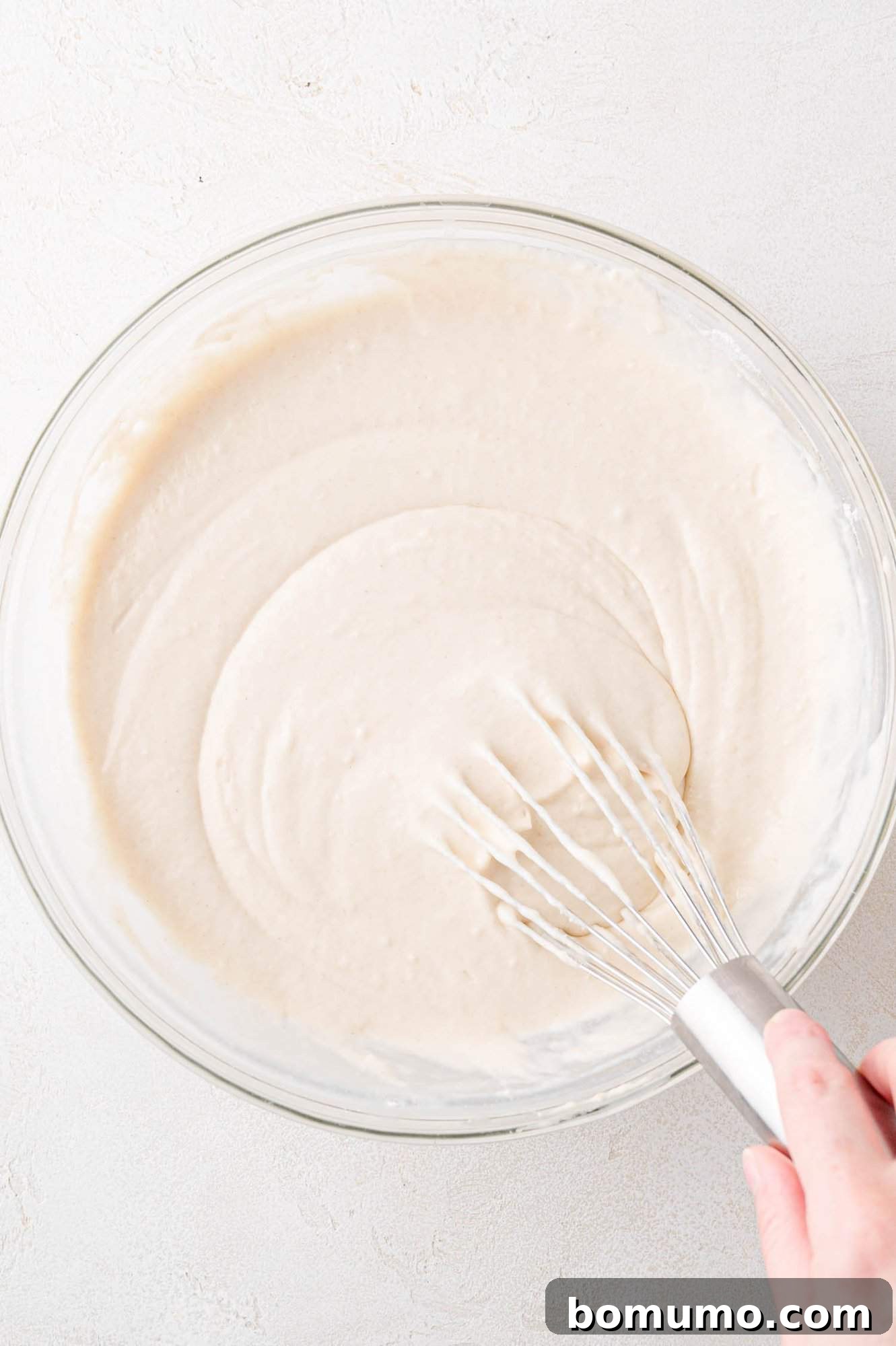 A hand gently whisks a creamy, light-colored pancake batter in a clear glass bowl on a pristine white surface, showcasing the smooth, consistent texture of the mixture.