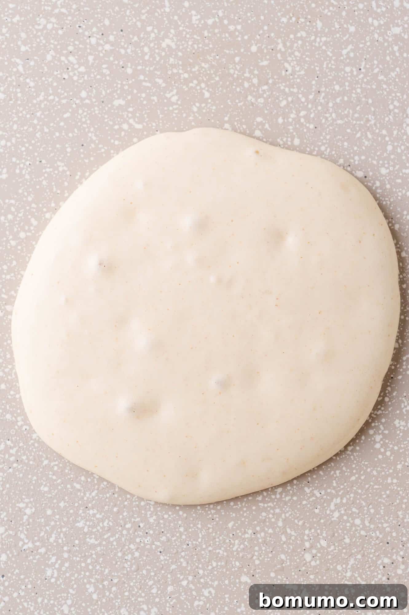 A perfectly round circle of light-colored, smooth pancake batter, adorned with small, active air bubbles, cooking on a dark, speckled griddle surface.