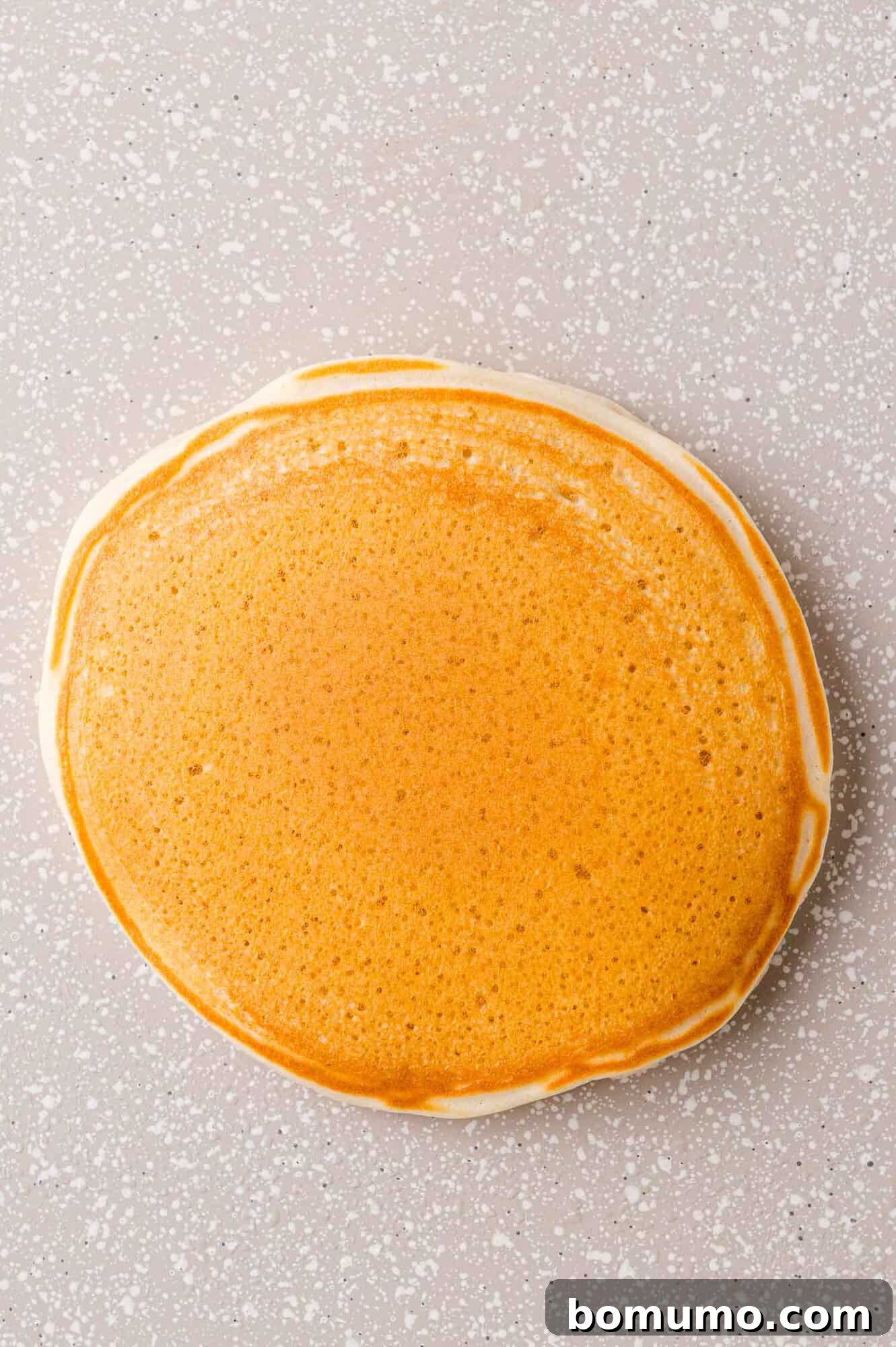 A single, beautifully golden-brown sweet cream pancake, perfectly cooked and ready to be served, resting on a dark, speckled gray griddle surface.