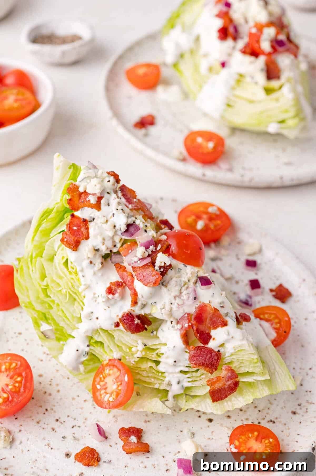 Two pristine wedge salads sit on speckled white plates, adorned with creamy blue cheese dressing, bright cherry tomatoes, finely diced red onions, and crispy bacon bits. One plate is prominently featured, while another is subtly visible in the background, creating an inviting culinary scene.