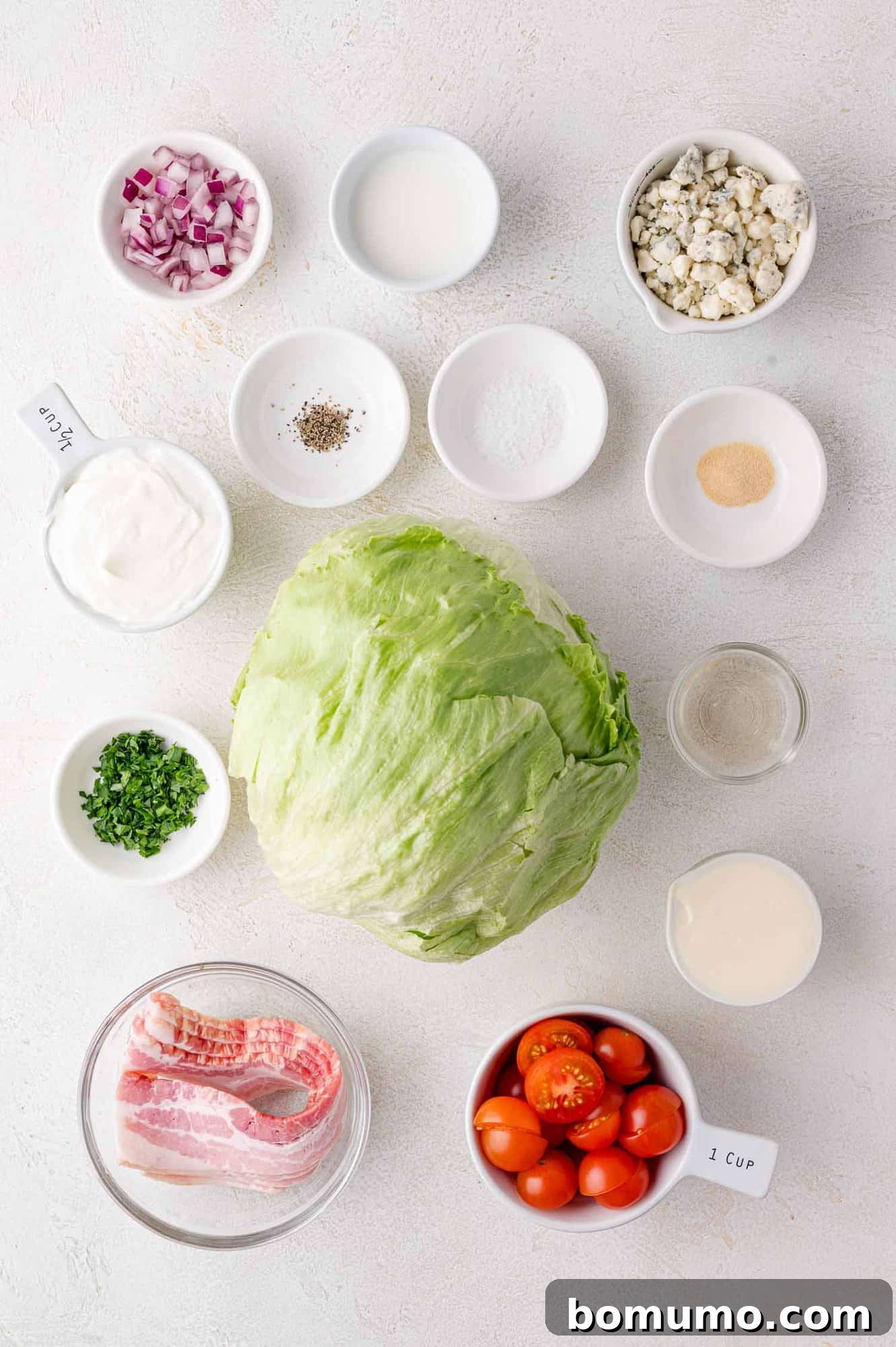 A collection of fresh ingredients for a wedge salad, including a crisp head of iceberg lettuce, vibrant cherry tomatoes, strips of bacon, red onion, a container of blue cheese, sour cream, fresh parsley, and various seasonings, all neatly arranged on a white surface.