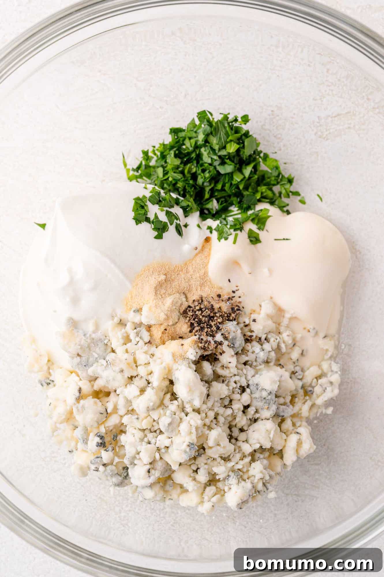 A glass bowl holds the initial ingredients for the blue cheese dressing, featuring creamy mayonnaise, crumbled blue cheese, garlic powder, black pepper, and fresh chopped parsley waiting to be mixed.