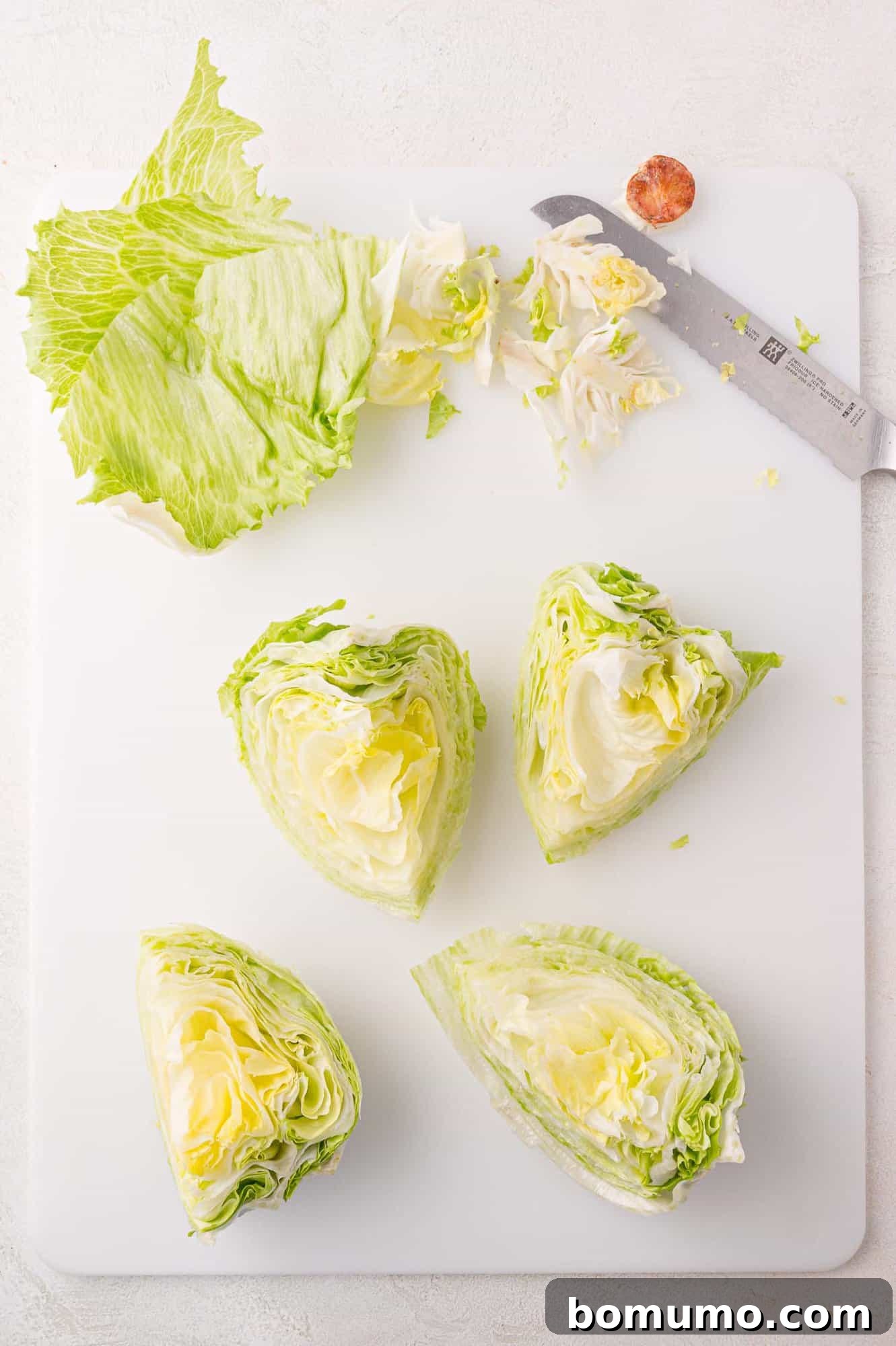 A perfectly cut head of iceberg lettuce sits on a cutting board, quartered with its core removed, alongside a chef's knife.