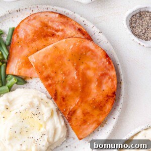 Two slices of glazed ham on a plate with mashed potatoes and green beans, seasoned with pepper.