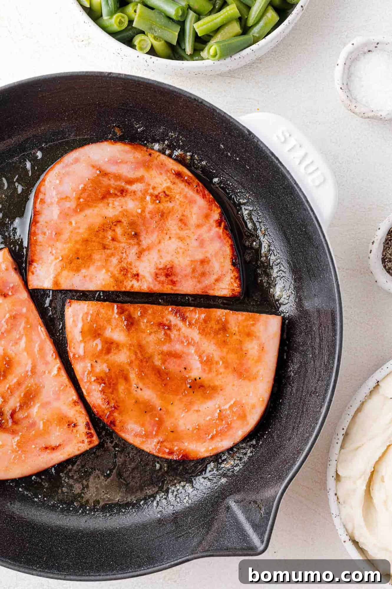 Slices of ham are sizzling in a skillet, their edges perfectly browned, with a vibrant side of green beans, a sprinkle of salt, and tender potatoes awaiting their turn, creating a mouth-watering culinary scene.