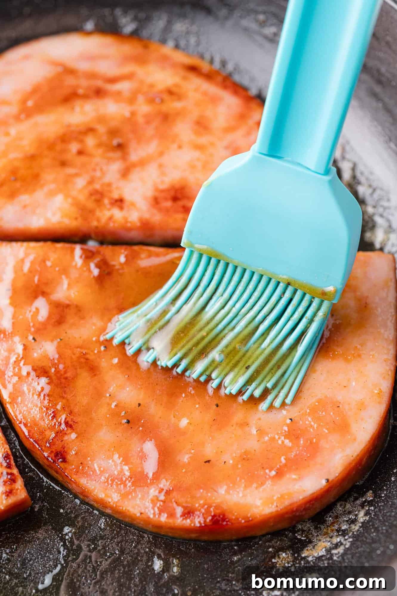 Two succulent slices of ham are being generously brushed with a rich, glossy glaze using a vibrant blue brush, all within the comforting warmth of a frying pan.