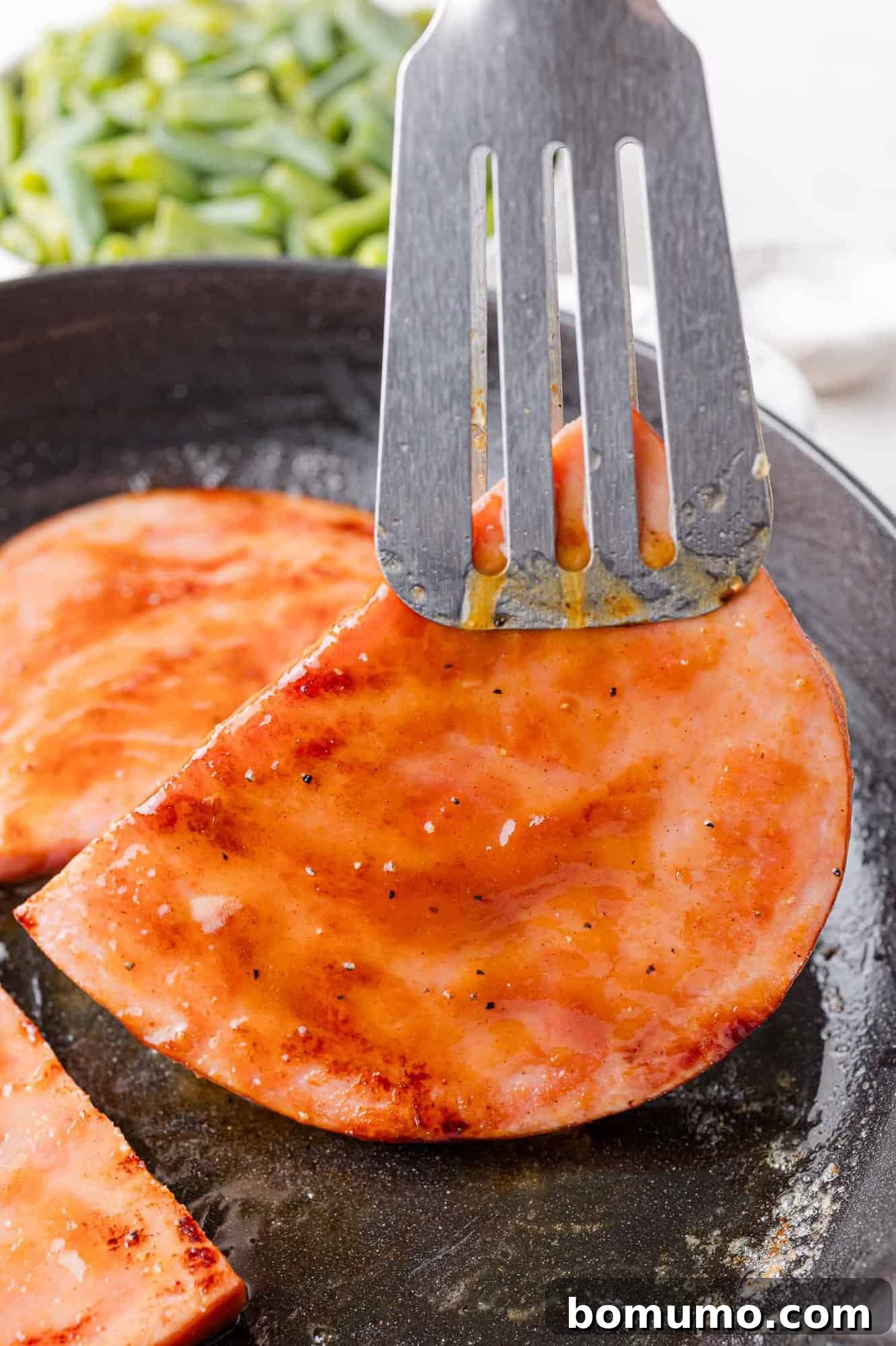 A spatula gently lifts a perfectly glazed slice of ham from a sizzling skillet, with a verdant bed of green beans blurred in the background, signaling a delicious meal in progress.