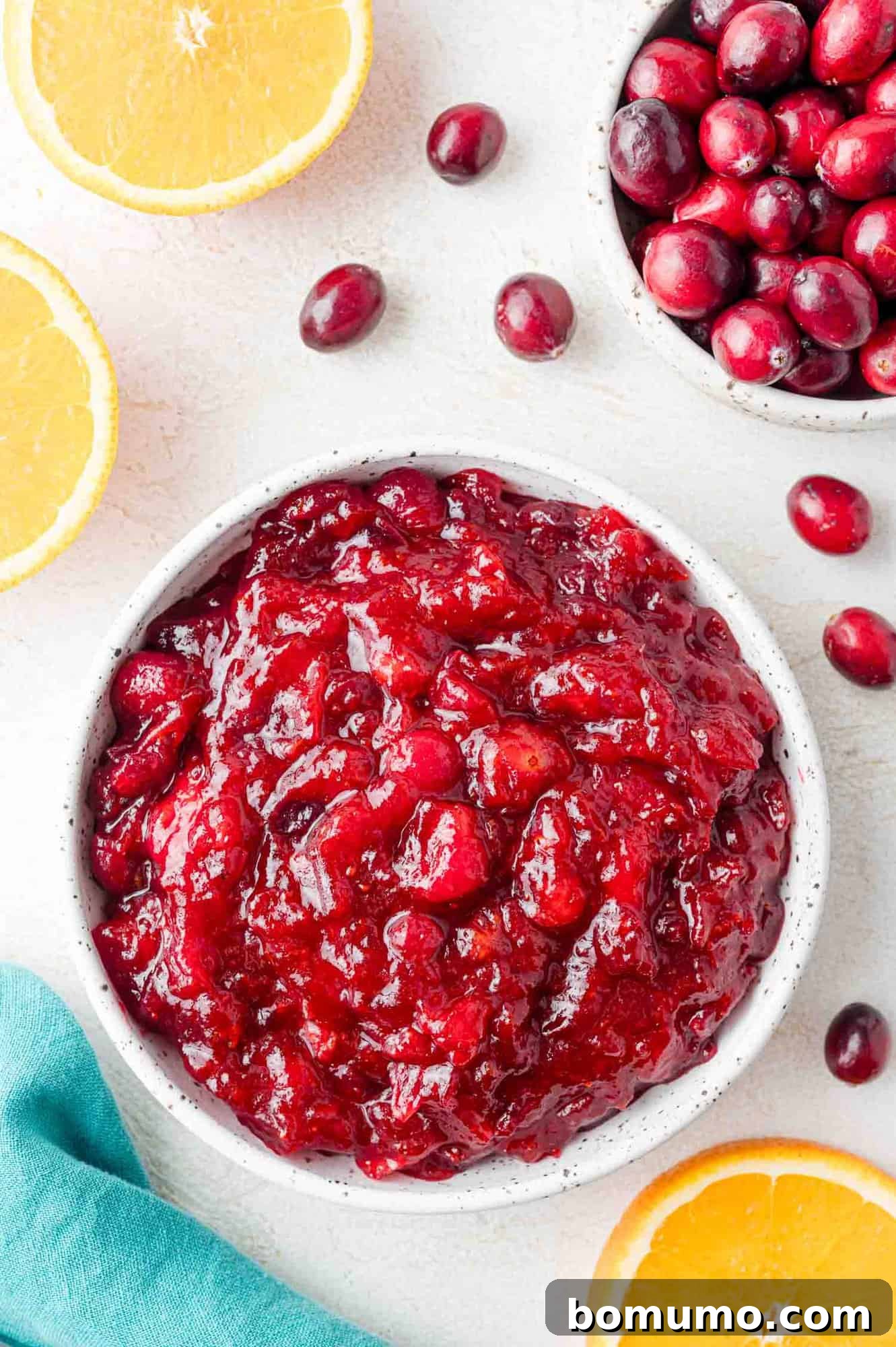 Vibrant Cranberry Orange Compote 2 Bowl of cranberry sauce surrounded by fresh cranberries and orange slices on a white surface, showcasing the vibrant colors and fresh ingredients.