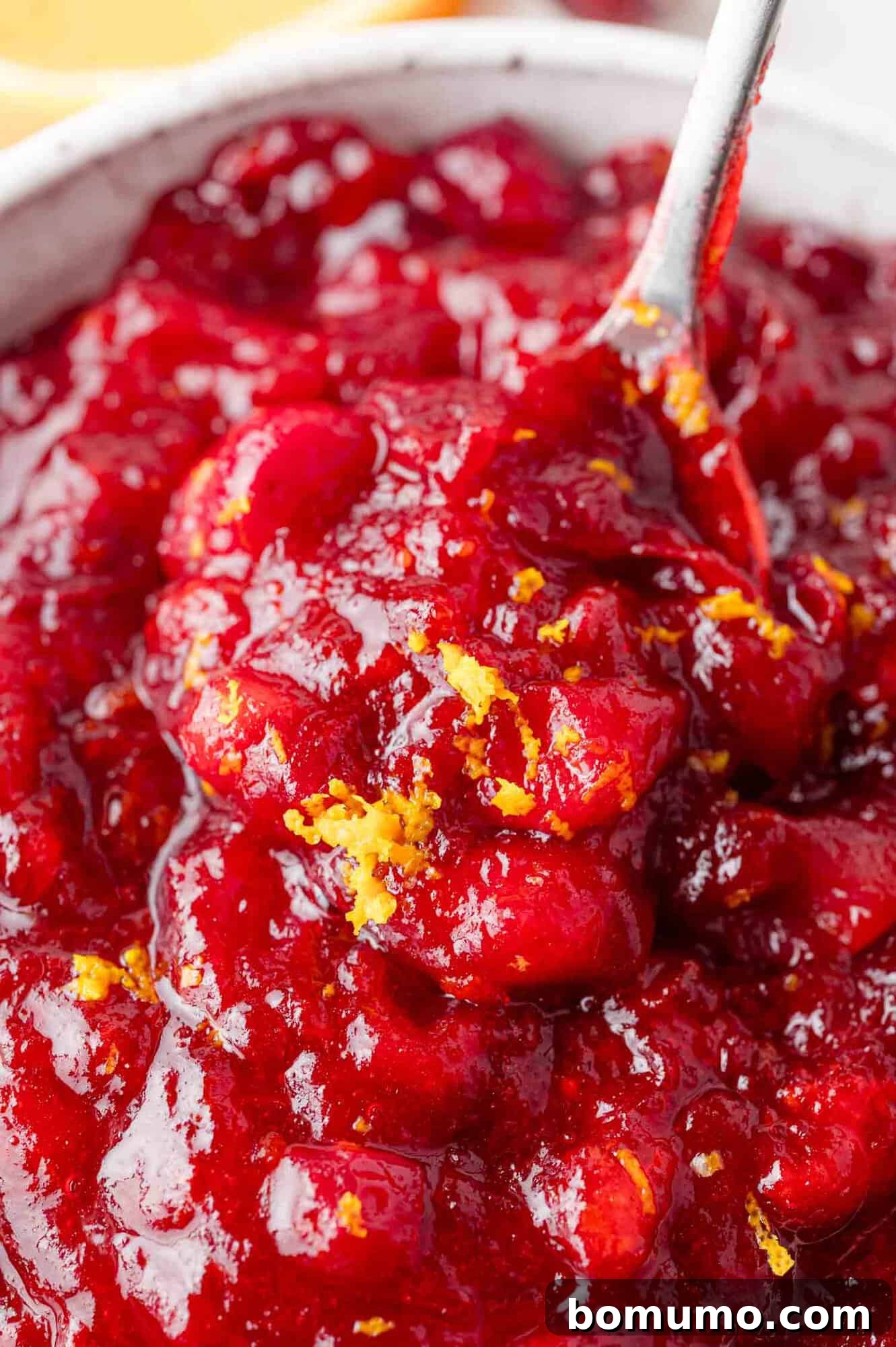 Vibrant Cranberry Orange Compote 3 Close-up of a bowl of cranberry sauce with visible orange zest on top, and a spoon partially submerged in the sauce, highlighting the texture and freshness.