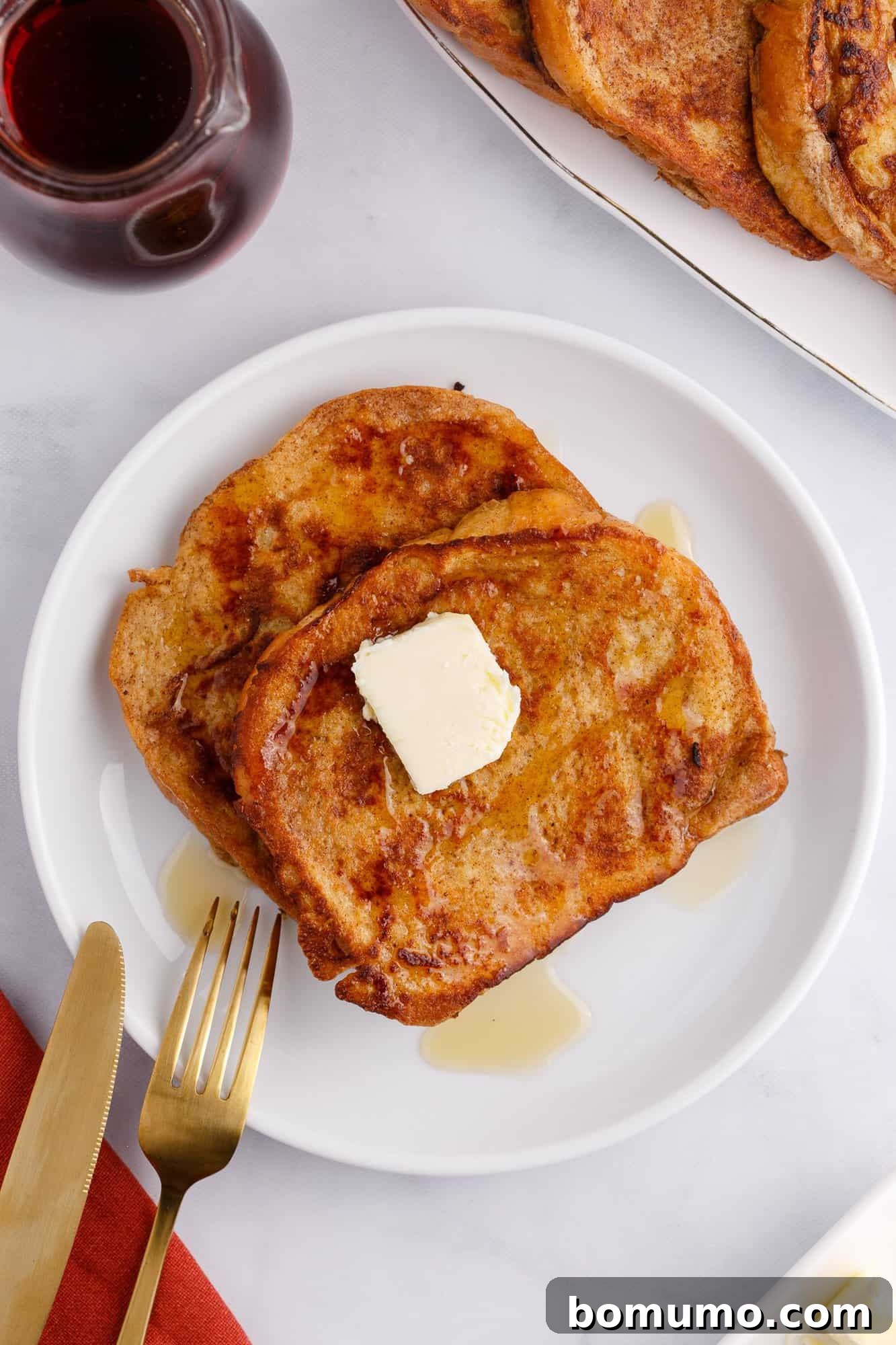 A plate of brioche French toast with a pat of butter and syrup, alongside a fork and knife on a white table. A small jug of syrup is nearby.
