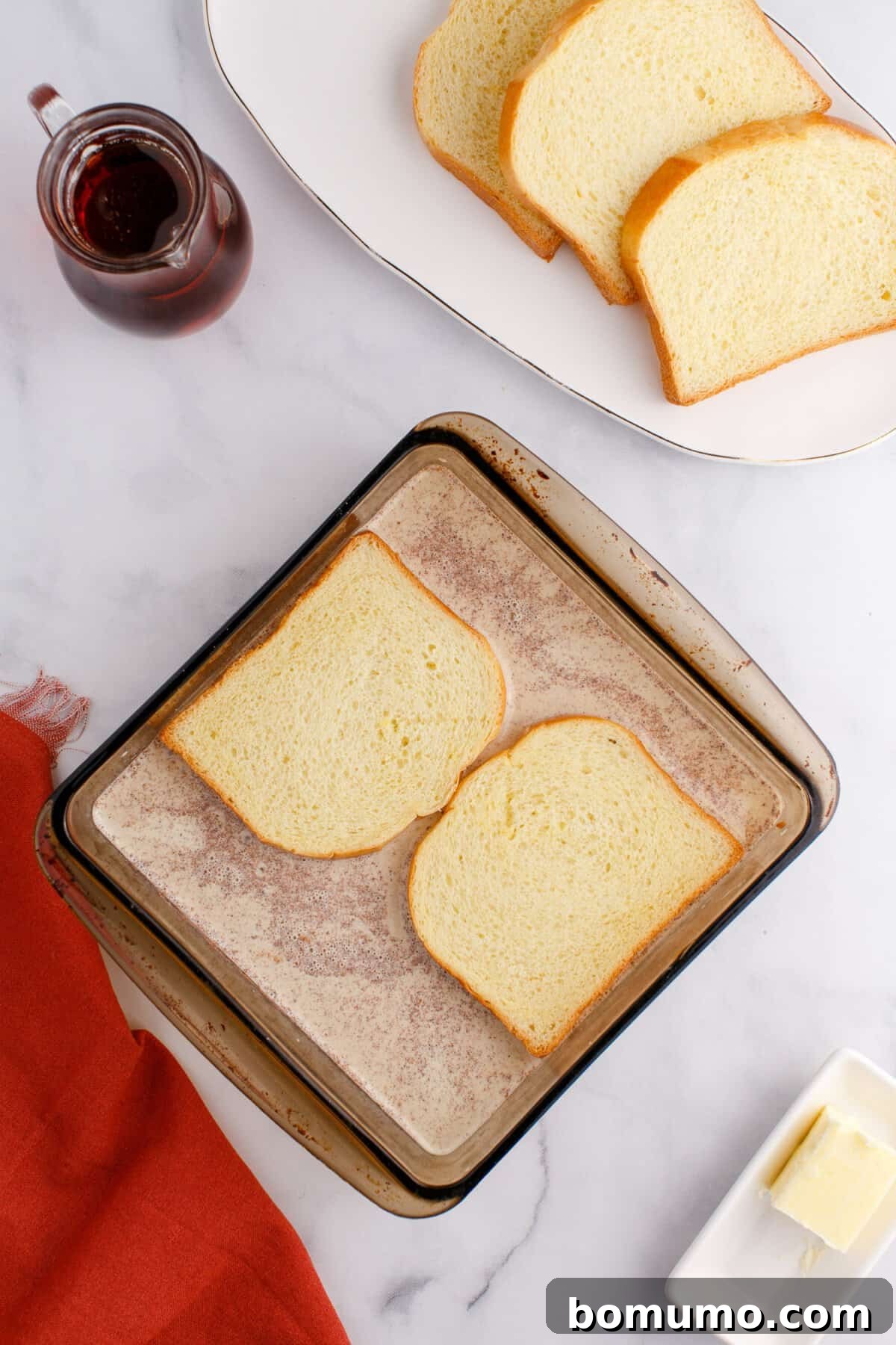 Two slices of bread in a glass baking dish, with additional slices on a plate, a small jug of syrup, a pat of butter, and a red cloth on a marble surface.