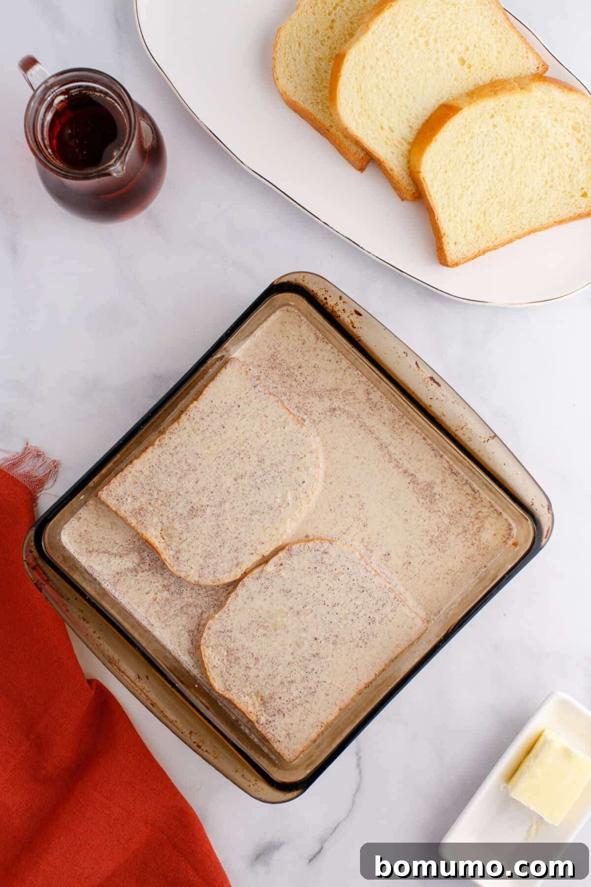 A glass baking dish with slices of bread soaked in a liquid mixture. Nearby are slices of bread, a small pitcher of syrup, a red napkin, and a butter dish on a marble surface.