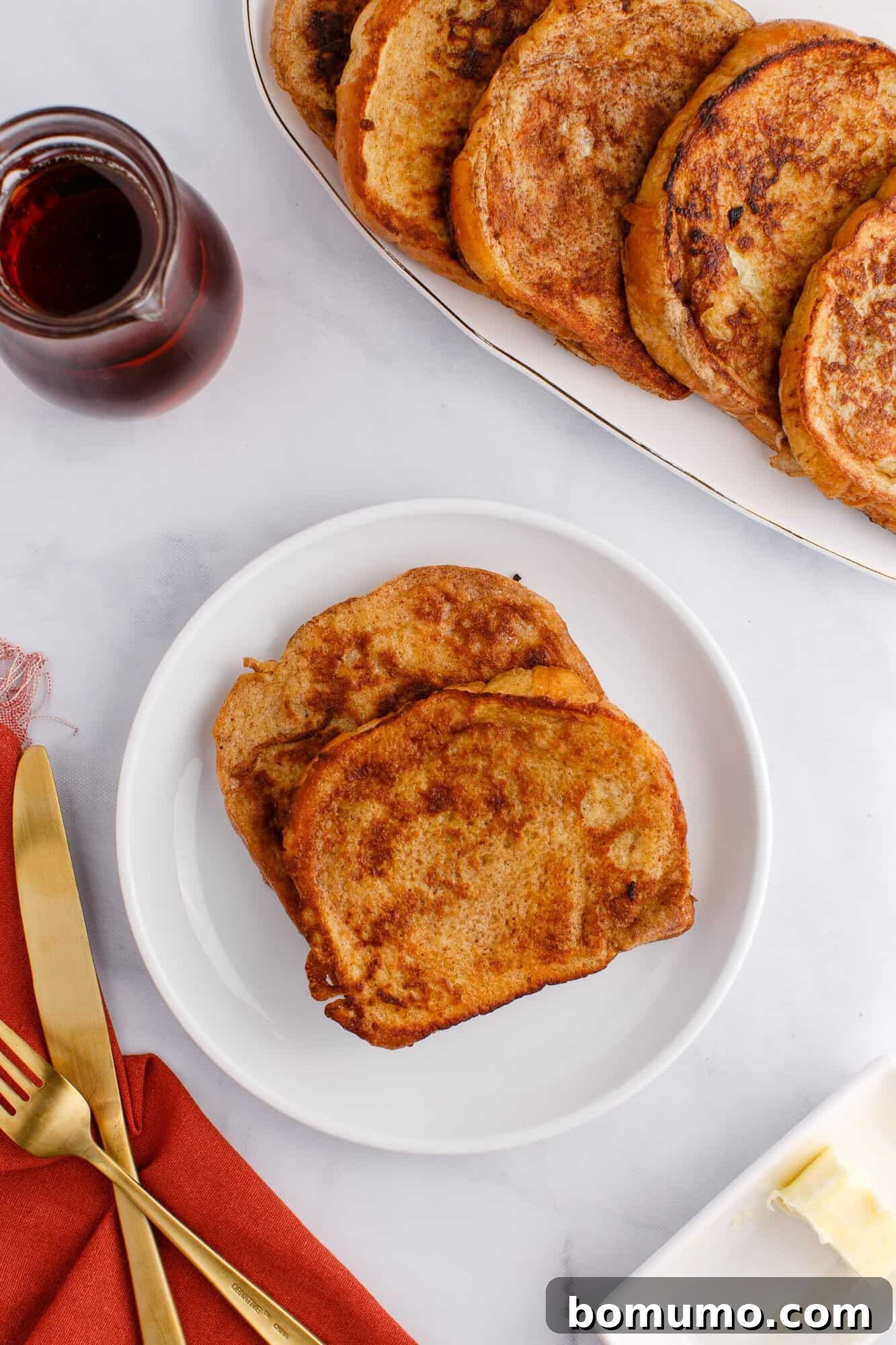 Two slices of French toast on a white plate, with a fork and knife beside a red napkin, a butter dish, and a small pitcher of syrup nearby.