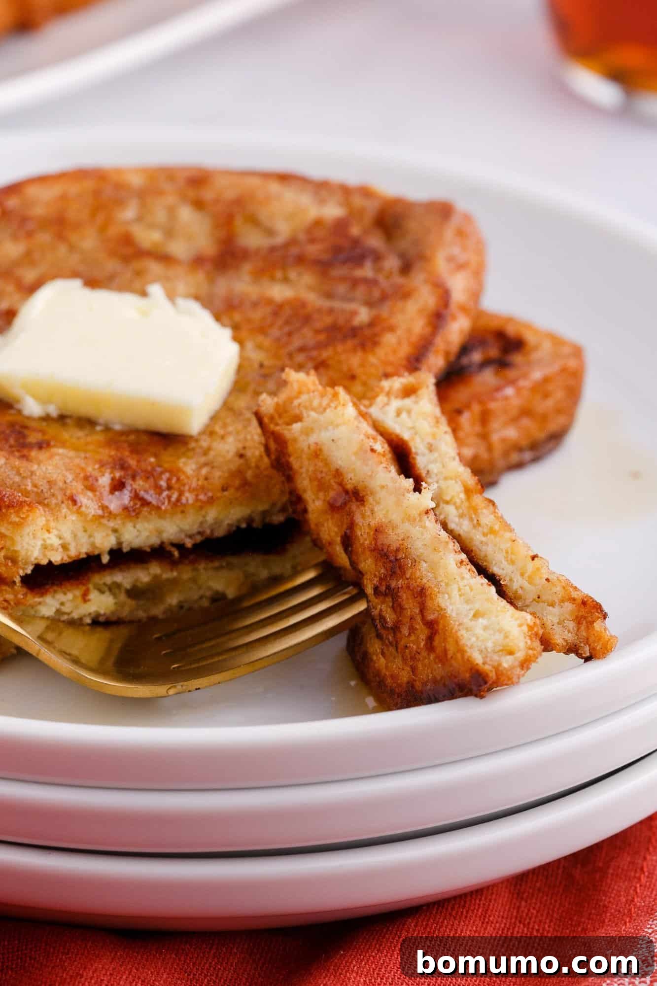 A stack of French toast topped with a butter pat sits on a white plate. A fork holds a cut piece of toast in front.