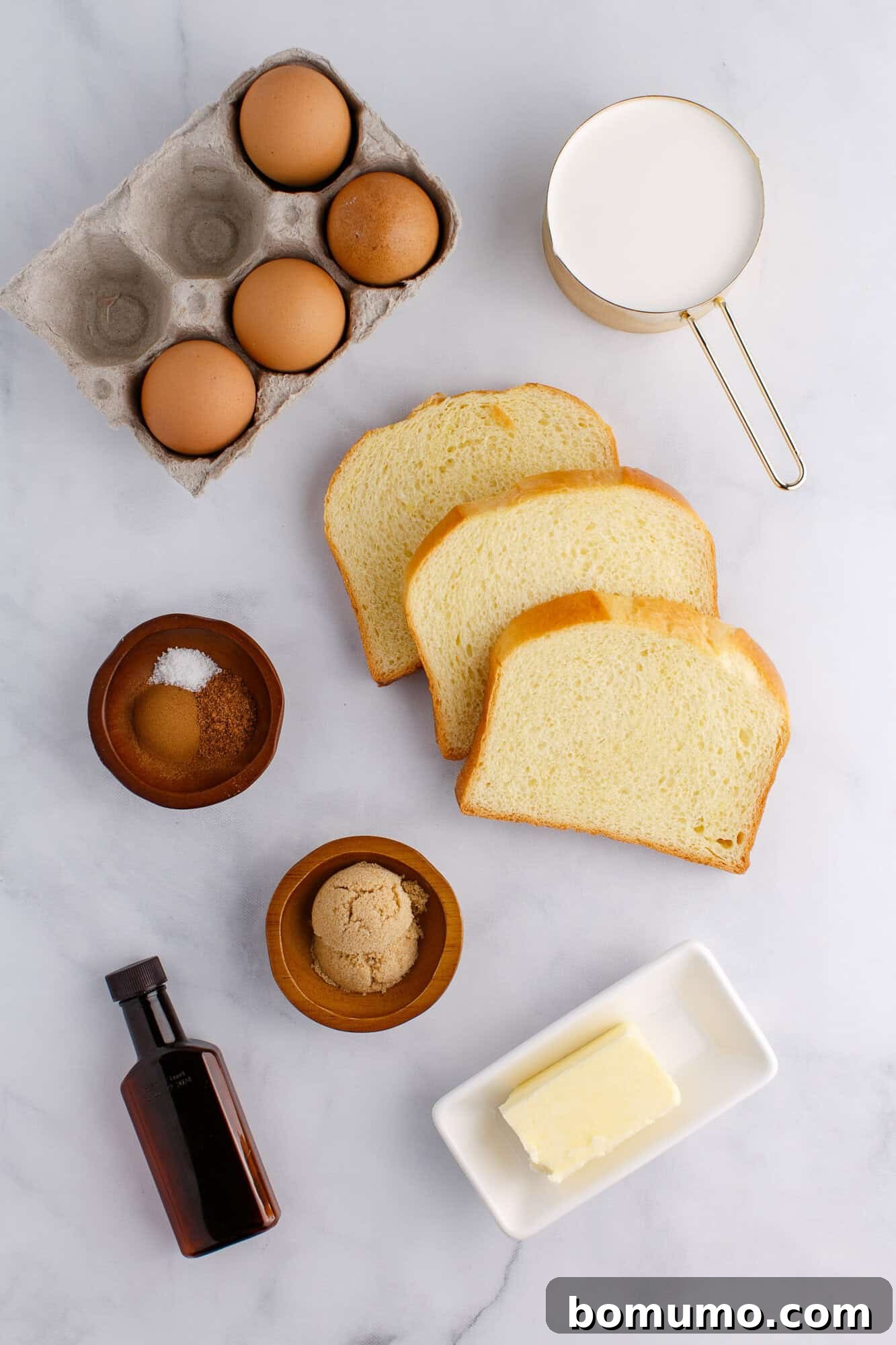 Ingredients for French toast: eggs in a carton, slices of bread, a cup of milk, butter, vanilla extract, brown sugar, and salt in bowls on a marble surface.