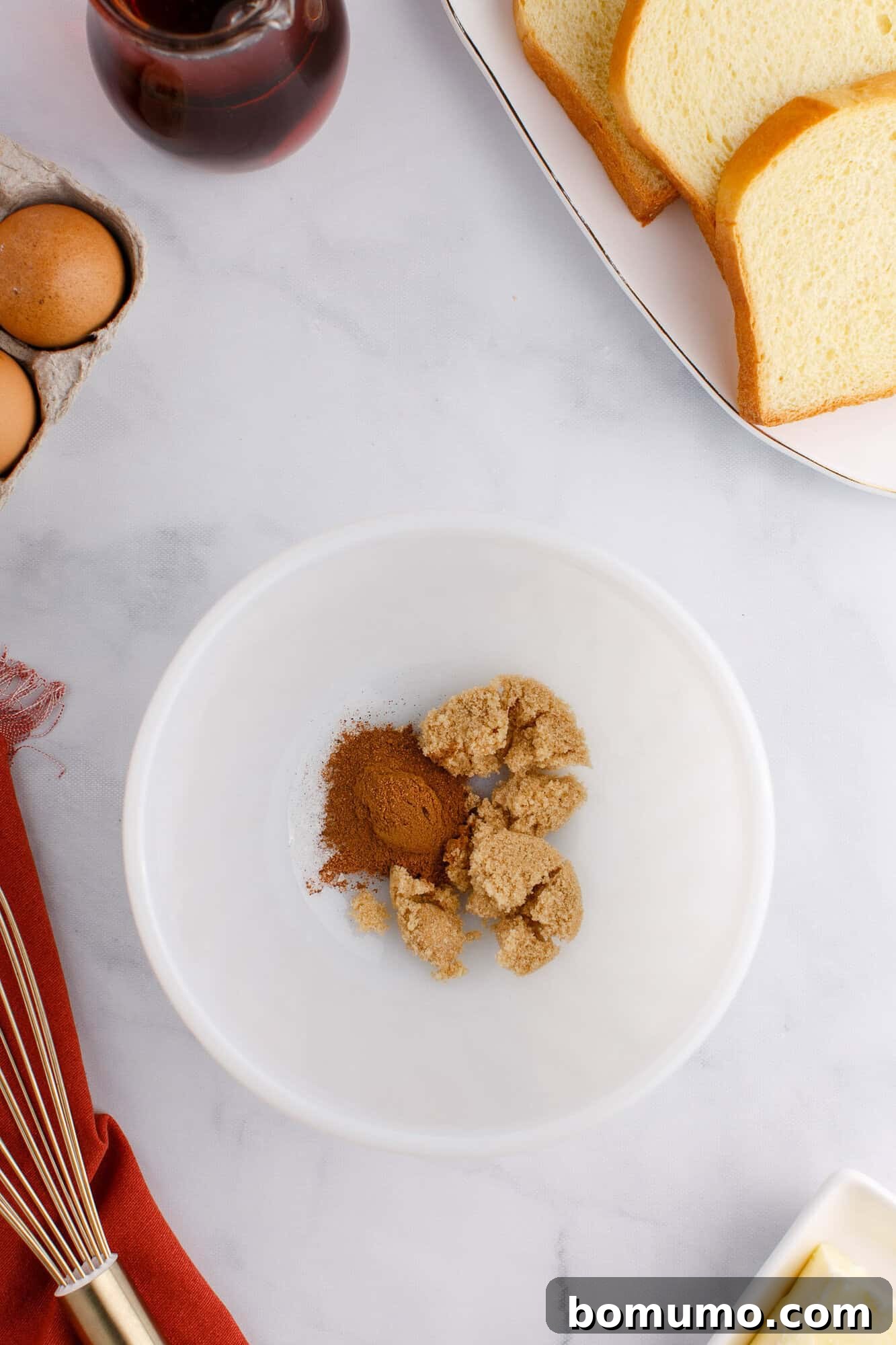 A bowl with brown sugar and cinnamon on a table with eggs, sliced bread, and a whisk nearby.