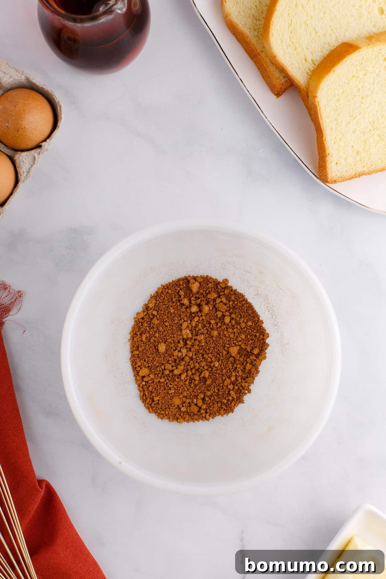 A white bowl with a mound of brown sugar on a countertop, surrounded by eggs, sliced bread.