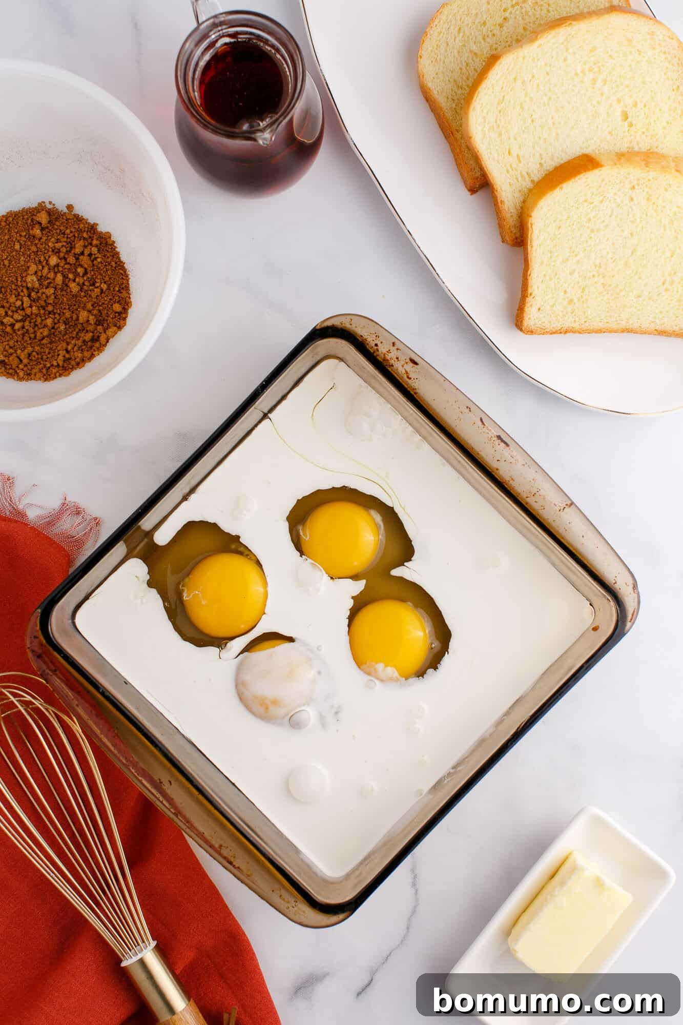 A baking dish with cream and three cracked eggs, slices of bread on a plate, and a small jar of syrup. A bowl of brown sugar and butter are nearby on a marble countertop.