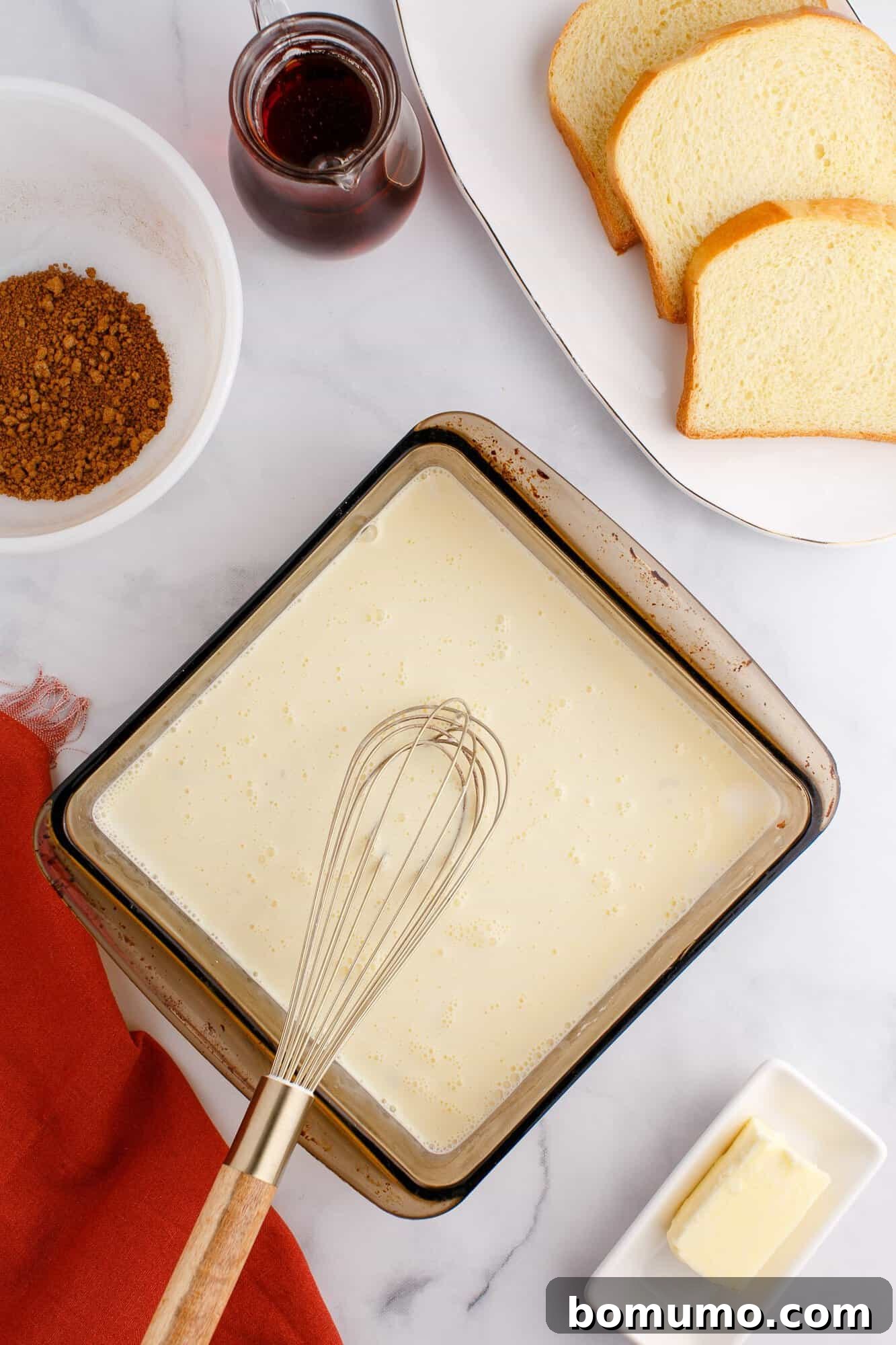 Ingredients for making French toast, including bread slices, a whisk in a bowl of batter, a bowl of spices, a dish of butter, and a jar of syrup on a marble surface.