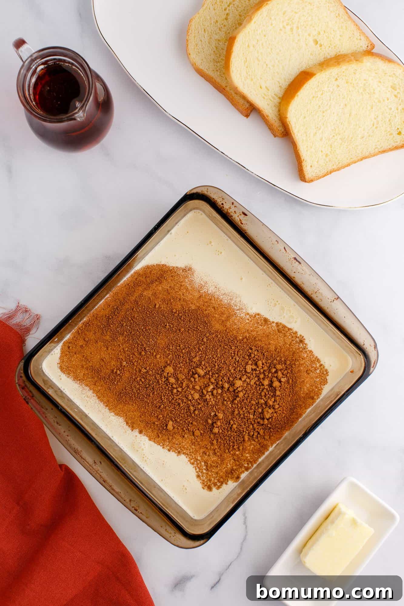 A baking dish with layered cream and brown sugar, alongside two slices of bread, a small pitcher of syrup, and a dish with a butter stick on a marble surface.