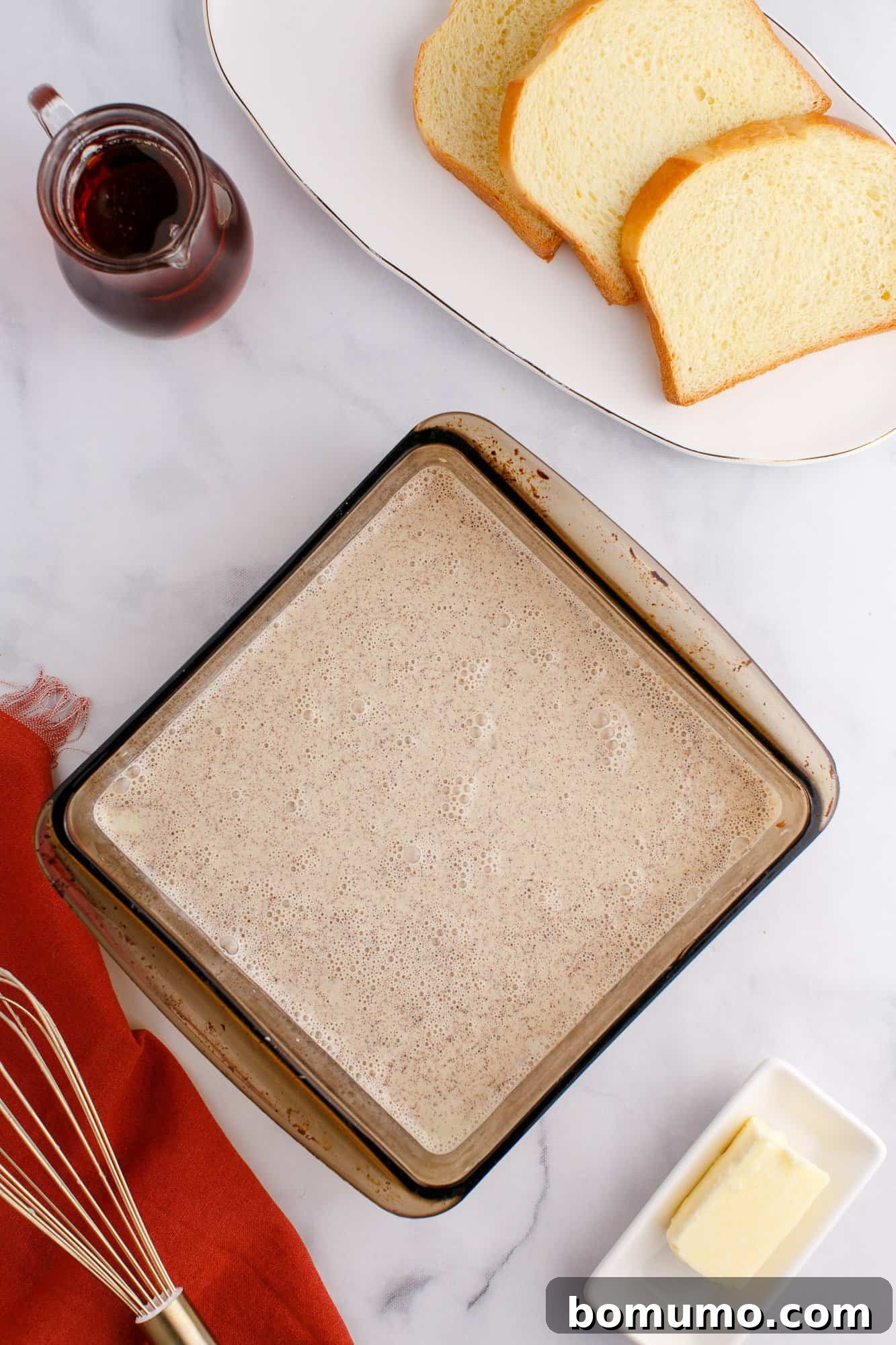 A glass baking dish with a light brown mixture, a plate with sliced bread, a small pitcher of liquid, a stick of butter, a whisk, and an orange cloth on a marble surface.