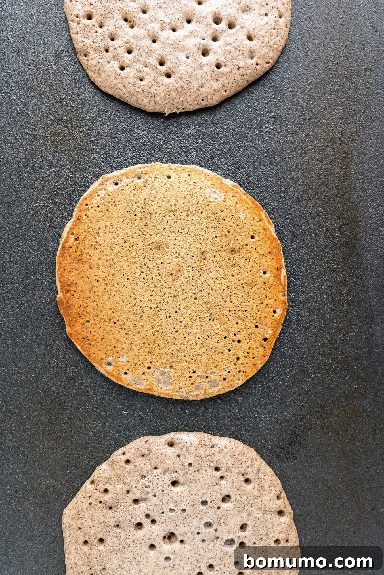 One buckwheat pancake expertly flipped to reveal a golden-brown underside, while two others still cook on the griddle, waiting their turn.