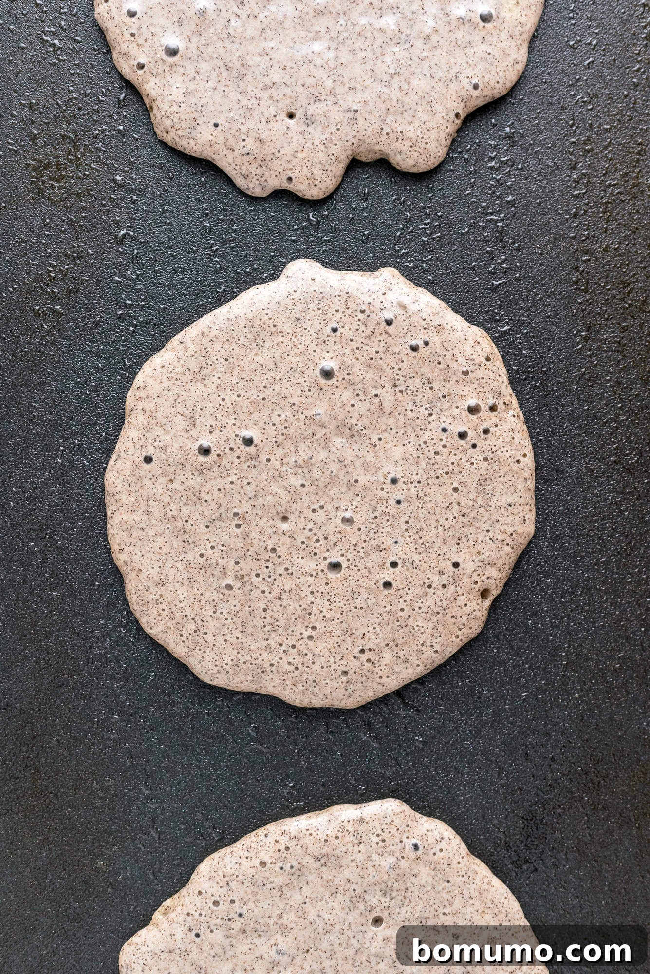 A batch of uncooked buckwheat pancakes gently cooking on a sleek black griddle, bubbles just beginning to form.
