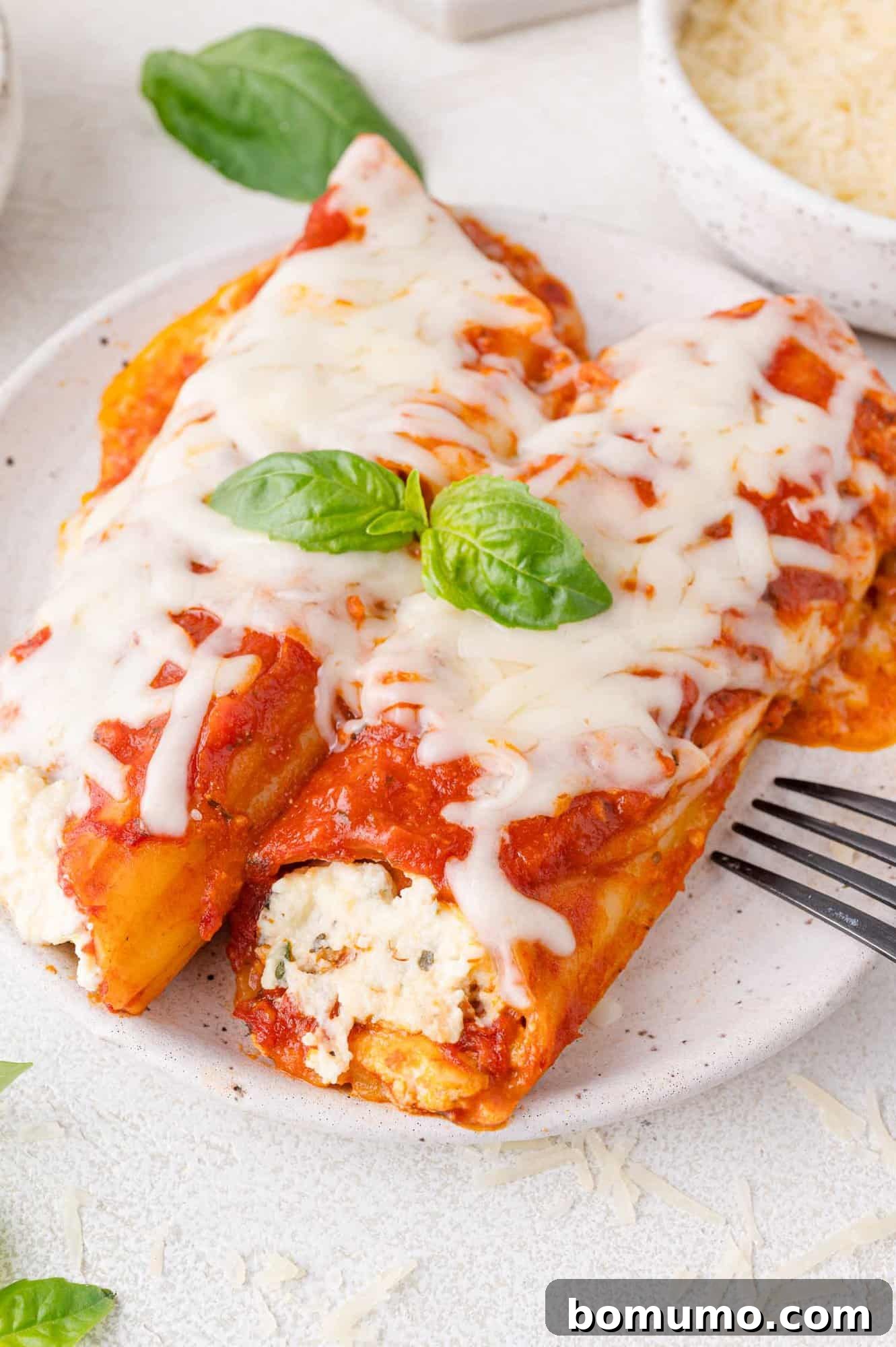 Two manicotti pasta tubes filled with ricotta cheese, covered in tomato sauce and melted cheese, garnished with basil leaves on a white plate. A fork rests beside the dish.