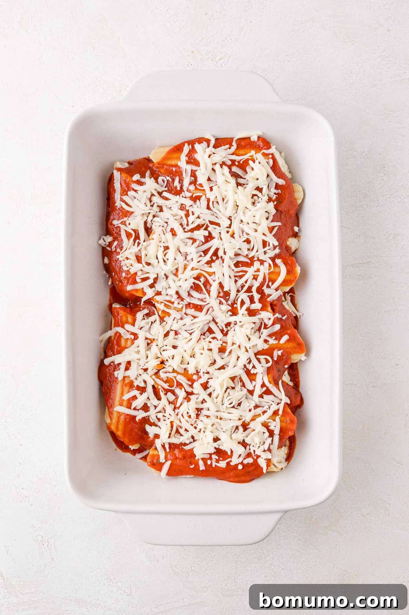A white baking dish filled with cannelloni topped with tomato sauce and shredded cheese.