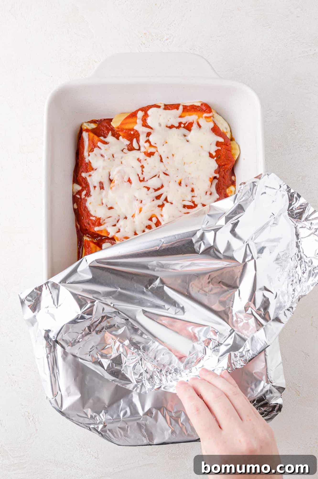A hand partially covers a baking dish with aluminum foil. The dish contains manicotti topped with tomato sauce and melted cheese.