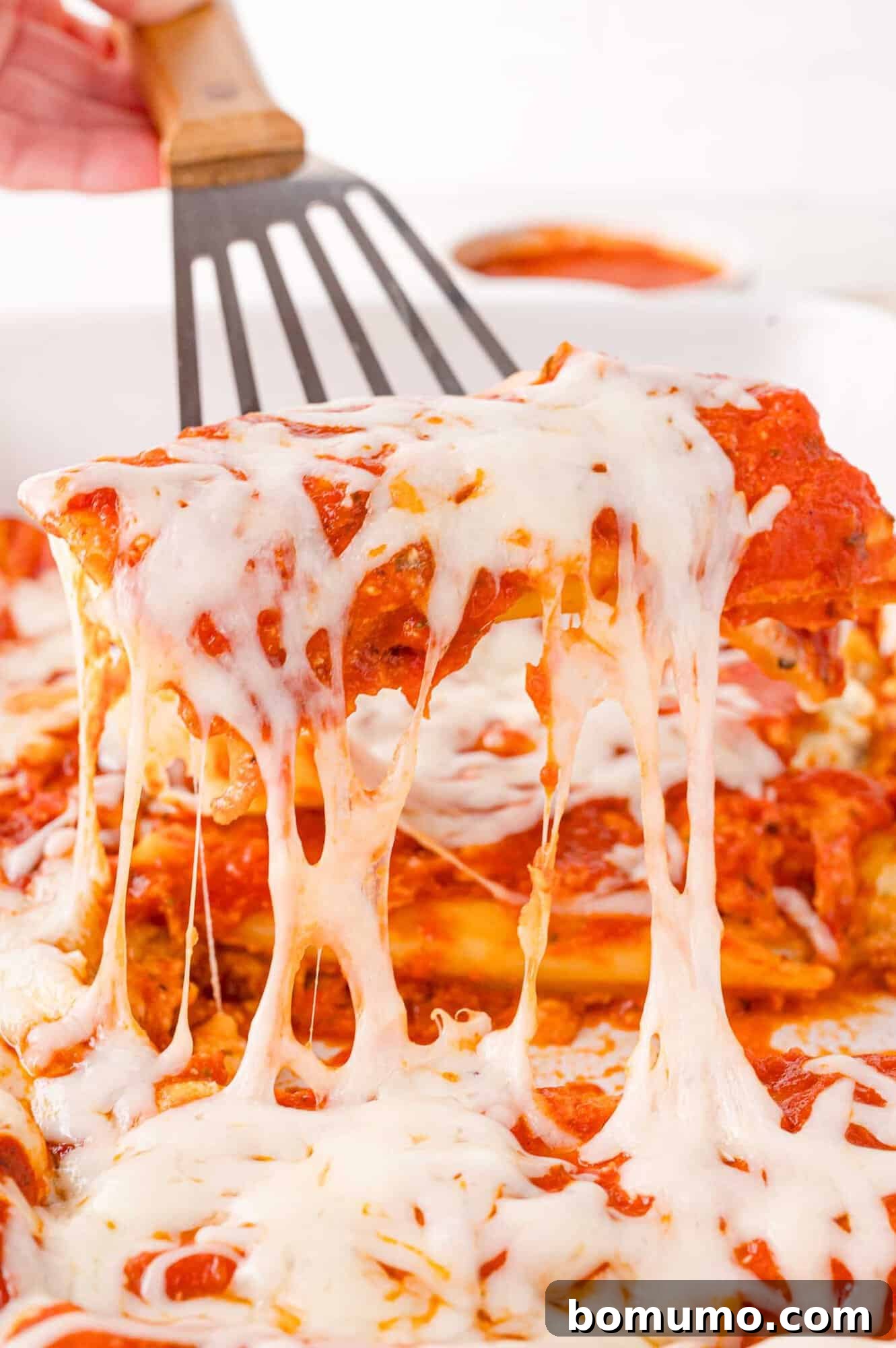A serving of cheesy manicotti is lifted from a dish with a spatula, showing melted cheese and red sauce.