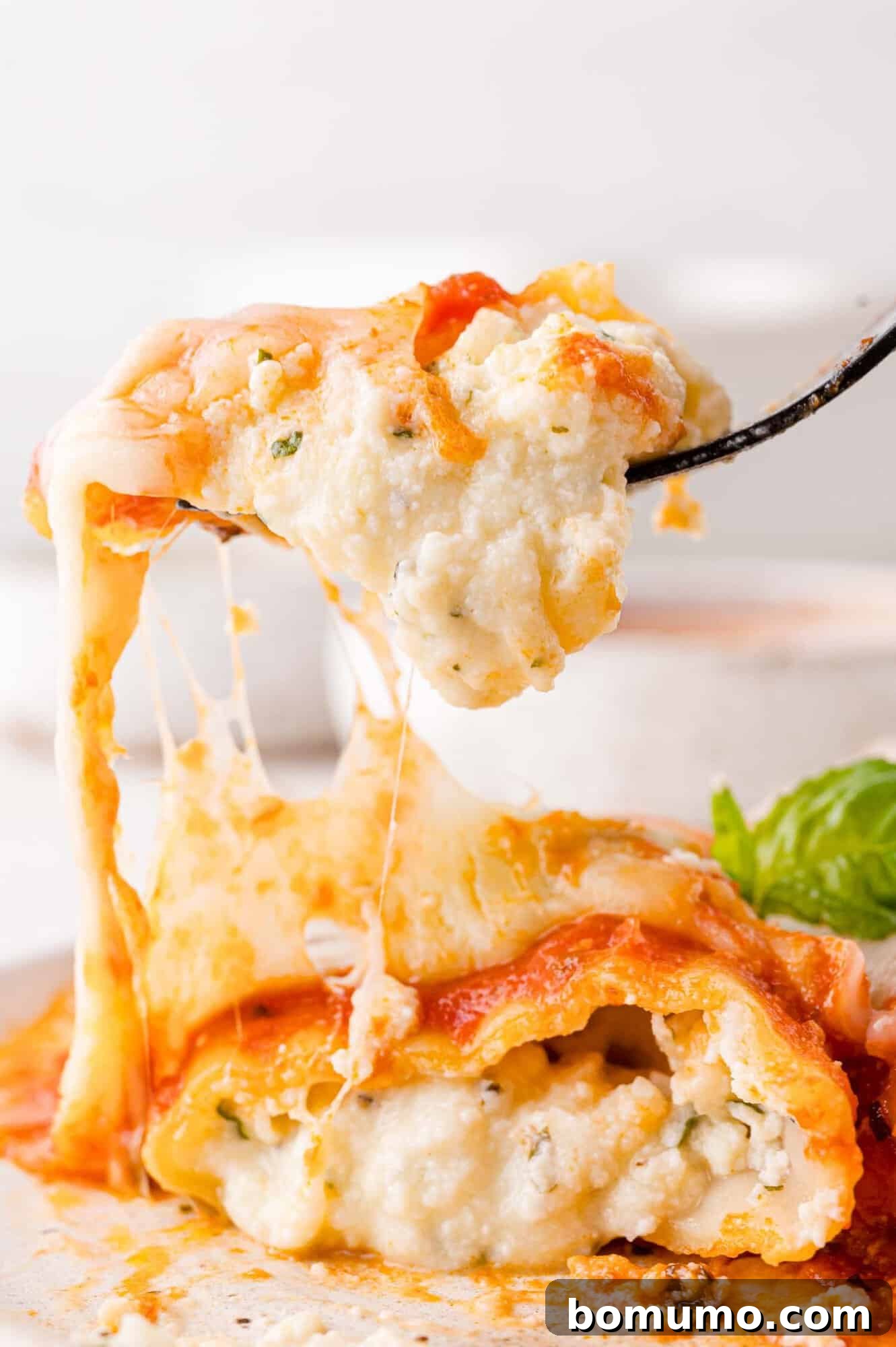 Close-up of a fork lifting a cheesy, ricotta-filled portion of manicotti from a plate, with visible layers of pasta, sauce, and basil garnish.
