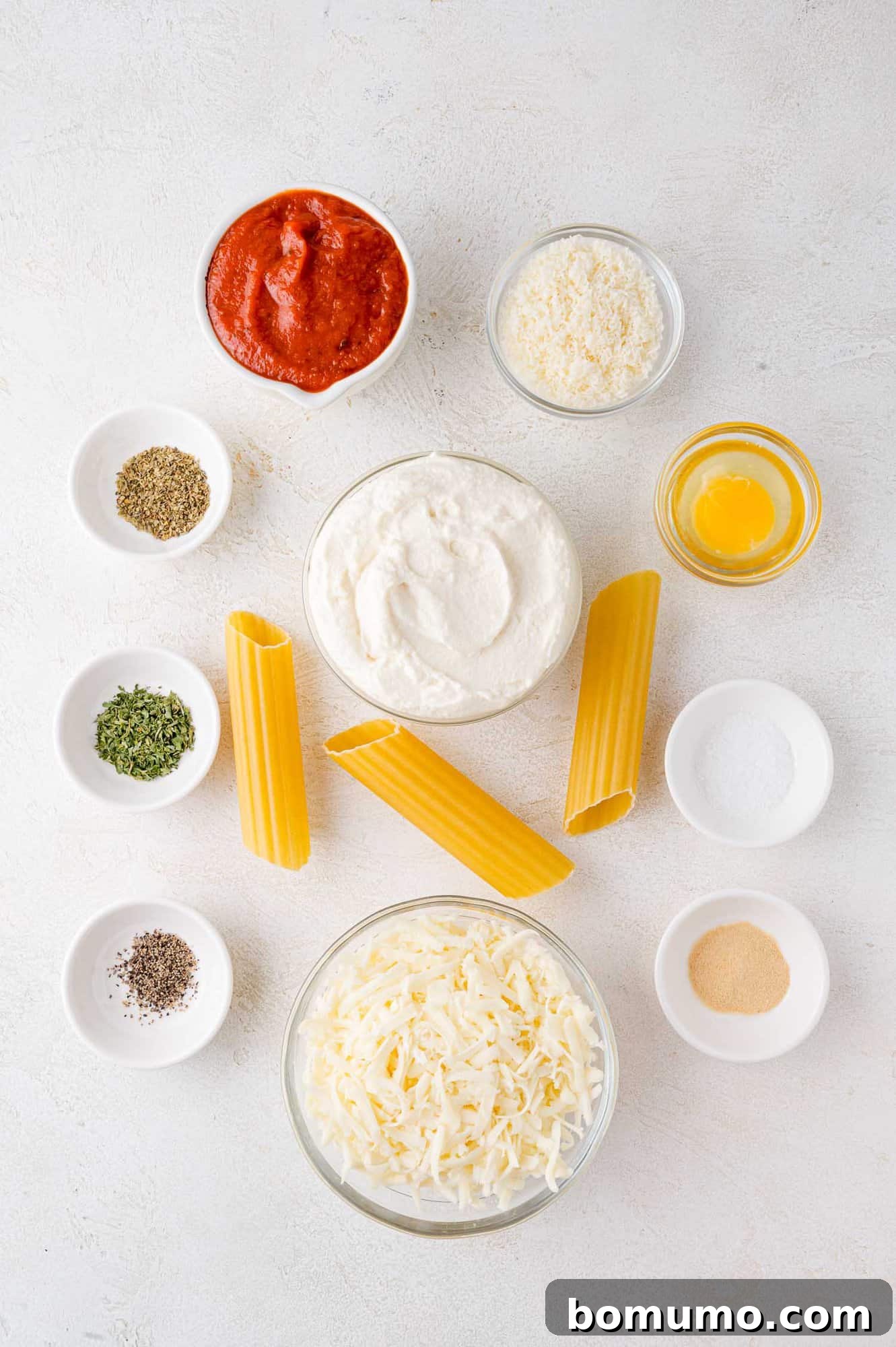 Ingredients for a pasta dish: ricotta, manicotti pasta, tomato sauce, shredded cheese, grated cheese, egg, spices, and seasonings arranged in bowls on a white surface.