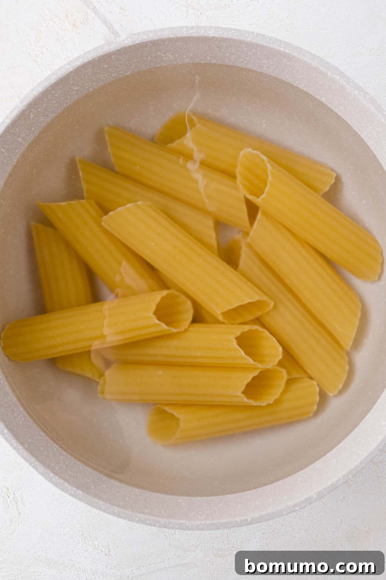 A white bowl filled with uncooked rigatoni pasta on a light surface.