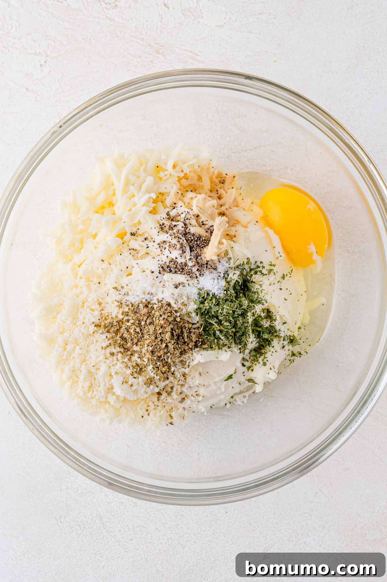 Clear glass bowl with shredded cheese, egg, herbs, pepper, and cream on a white surface.
