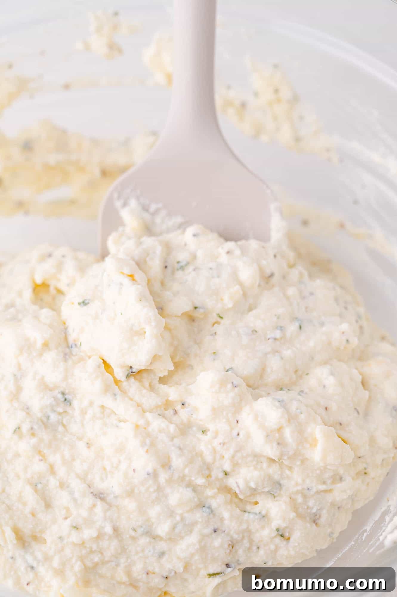 A white spatula in a bowl of creamy ricotta cheese mixture with herbs, viewed close-up.