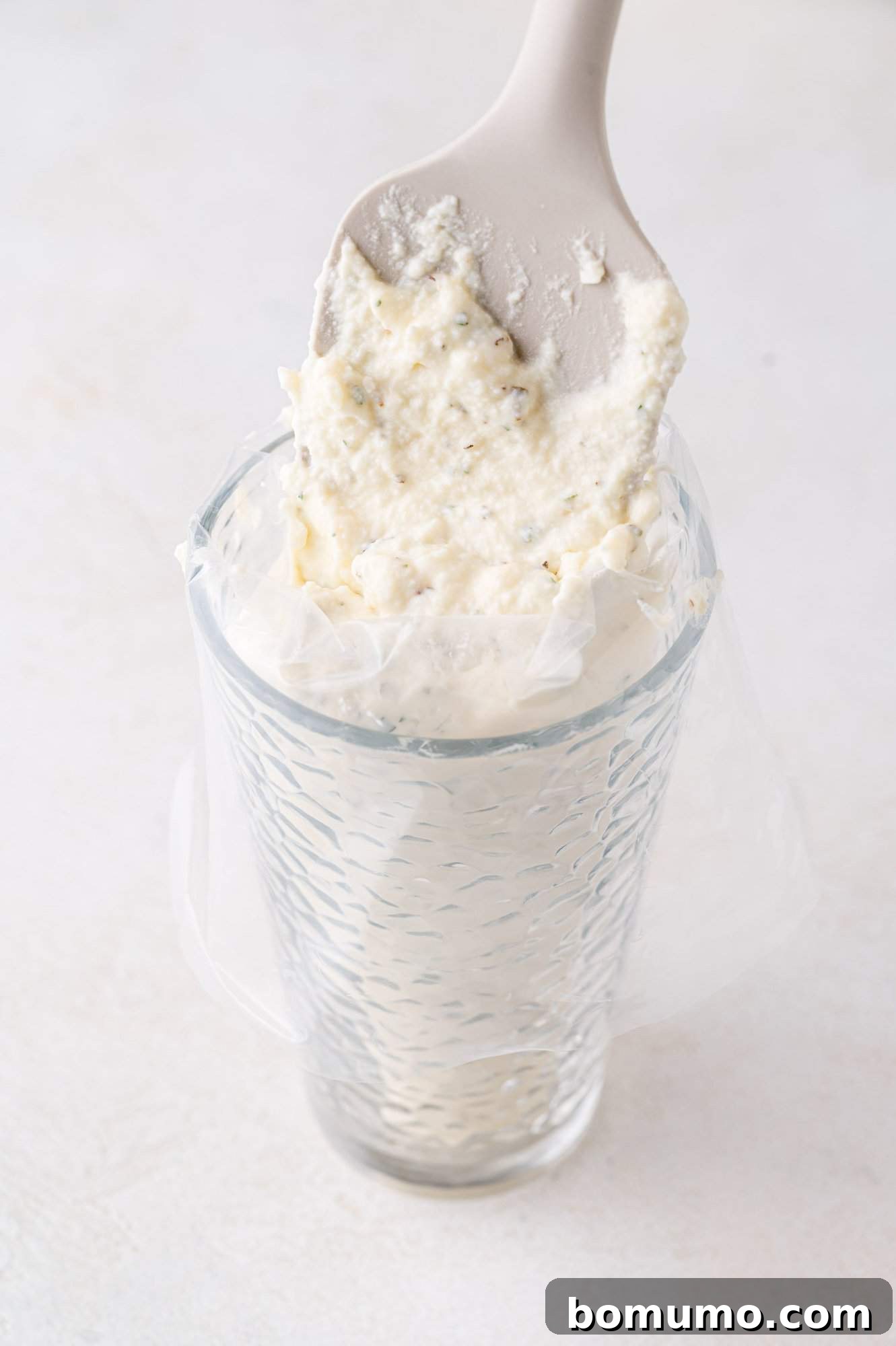 A spatula is scooping a white creamy mixture into a piping bag placed inside a textured glass on a light surface.