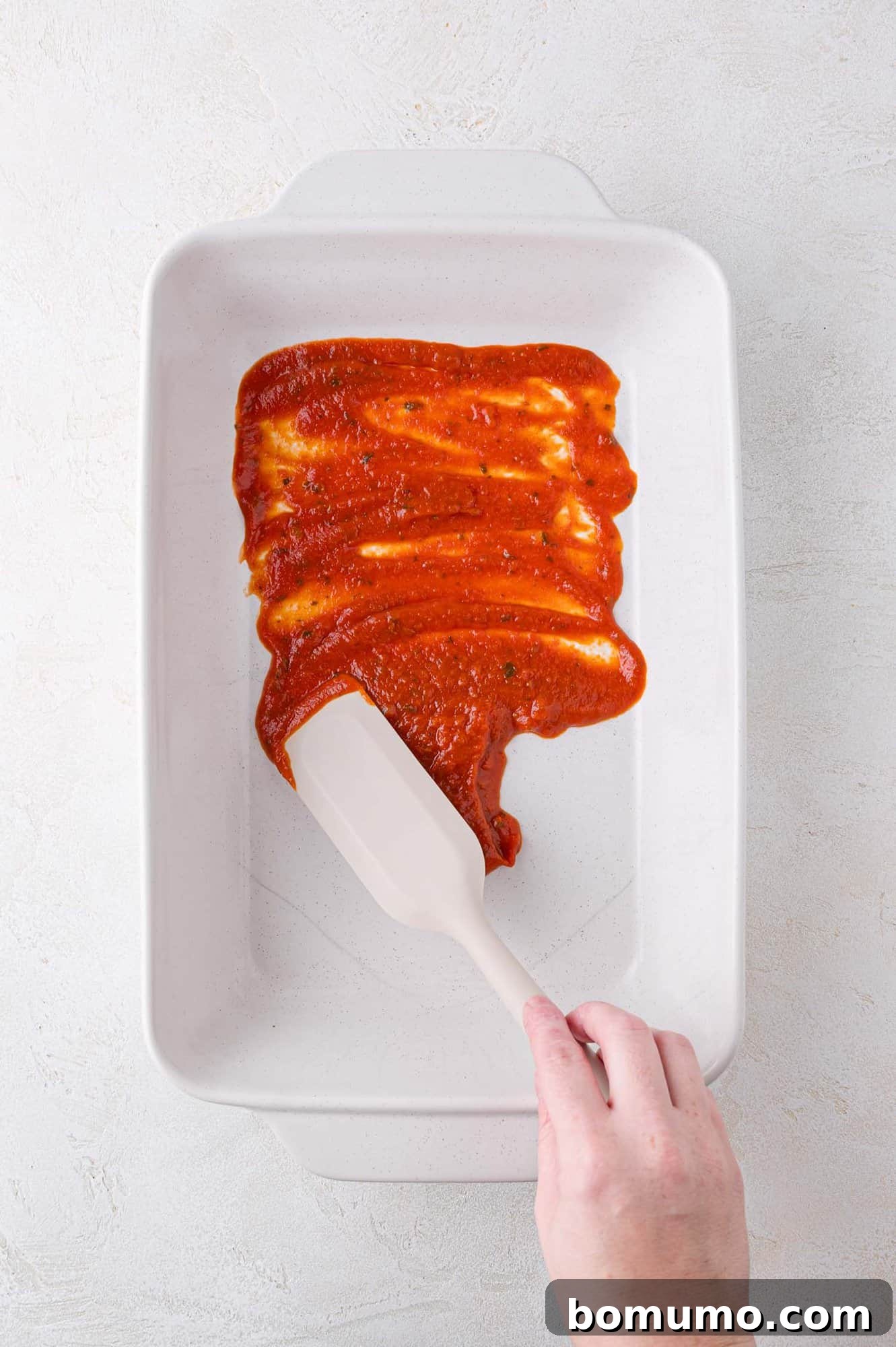 Hand spreading tomato sauce in a white rectangular baking dish with a spatula on a light background.